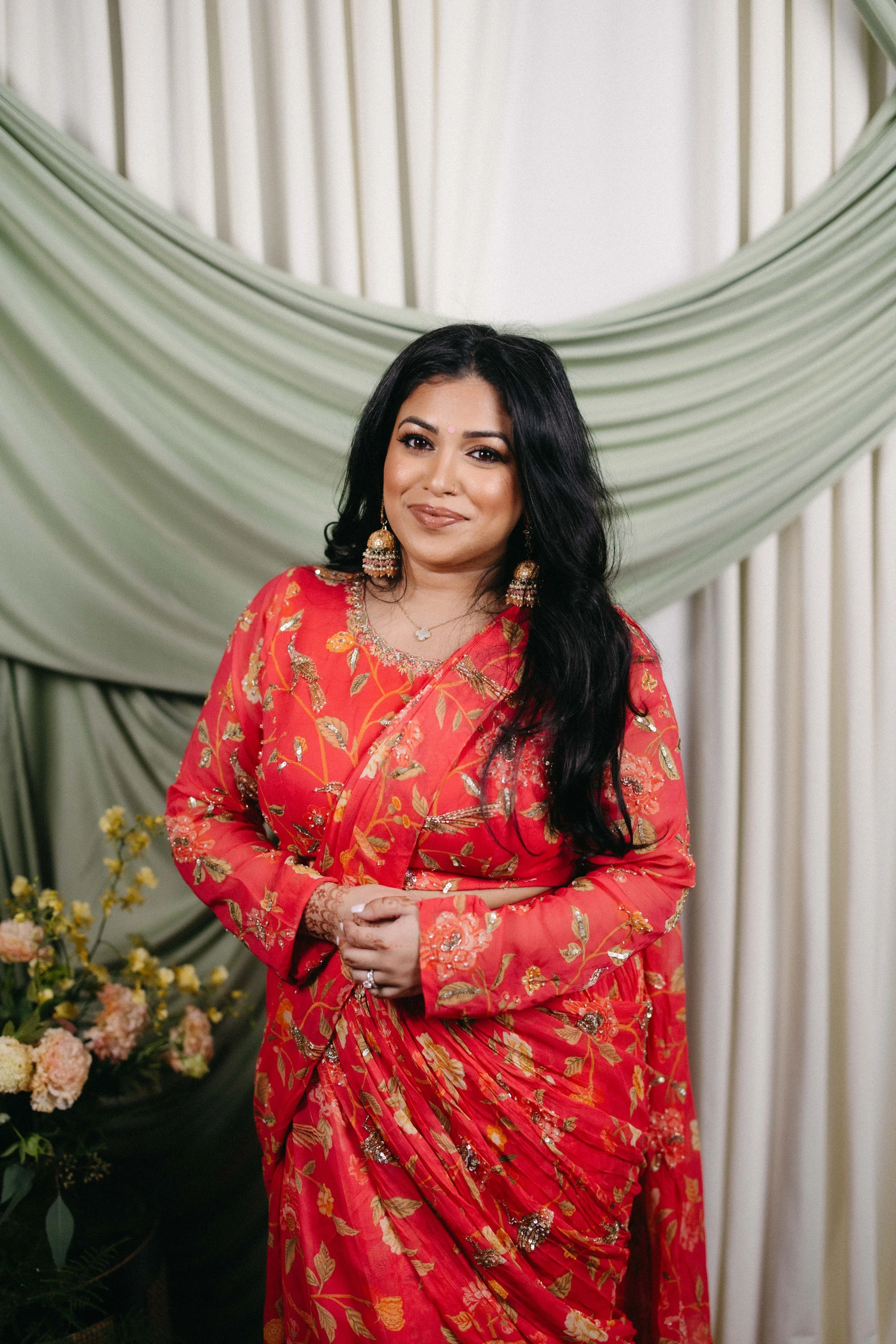 A woman with long black hair, wearing a red floral traditional outfit and jewelry, kneeling with hands clasped in front of a green draped backdrop with flowers on the side.