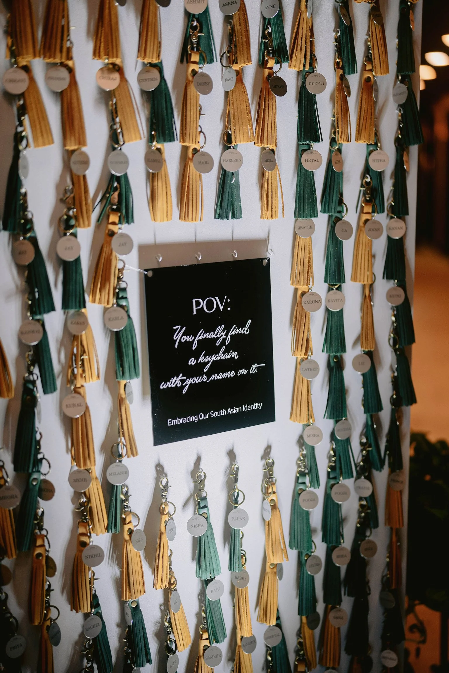 Multiple South Asian style keychains hanging on a wall, each with a round metal tag inscribed with names, attached to colorful tassels in gold and dark teal. A black sign in the middle reads: "POV: You finally find a keychain with your name on it. Em