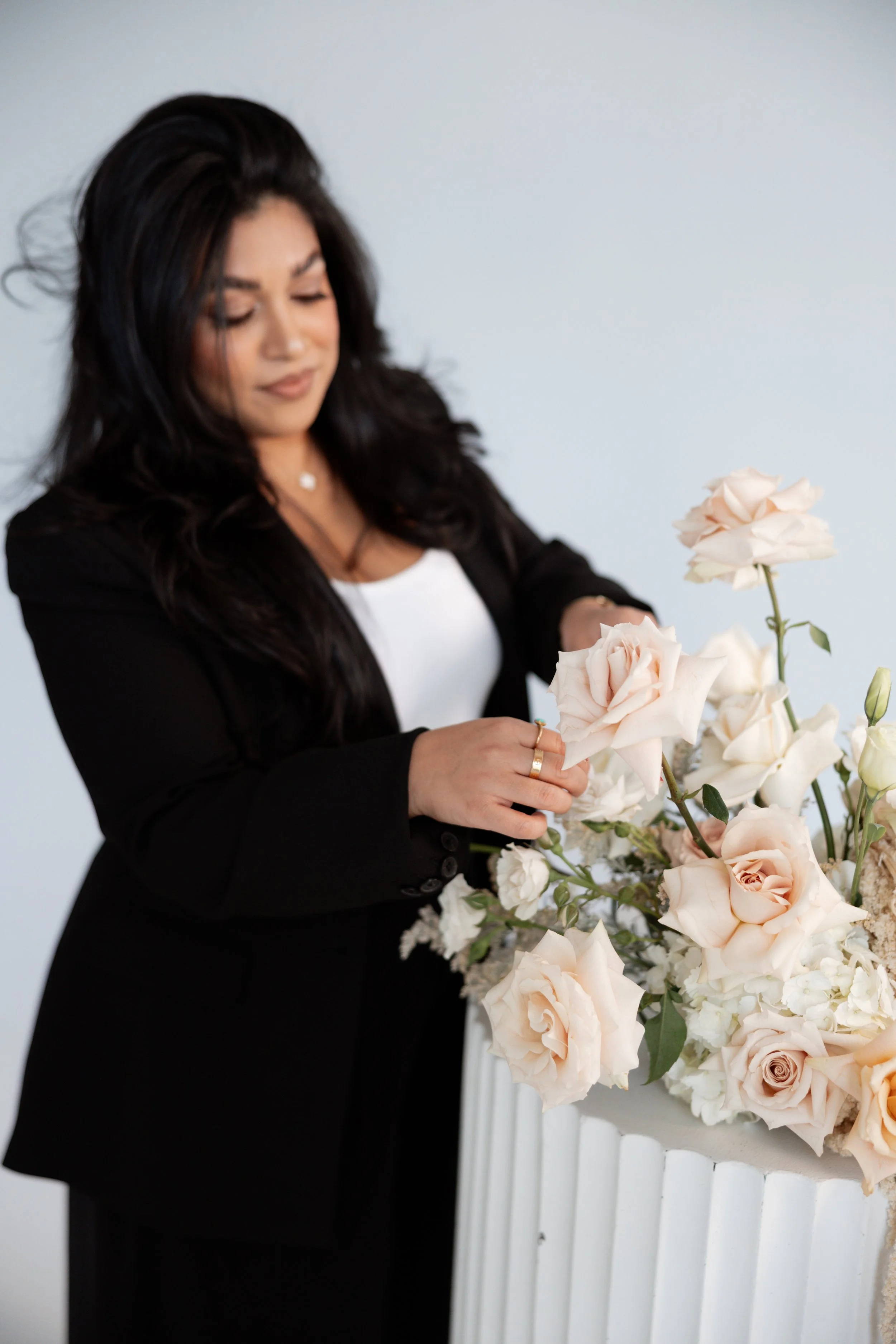 A woman with long black hair arranging pale pink roses in a white vase against a plain white background.