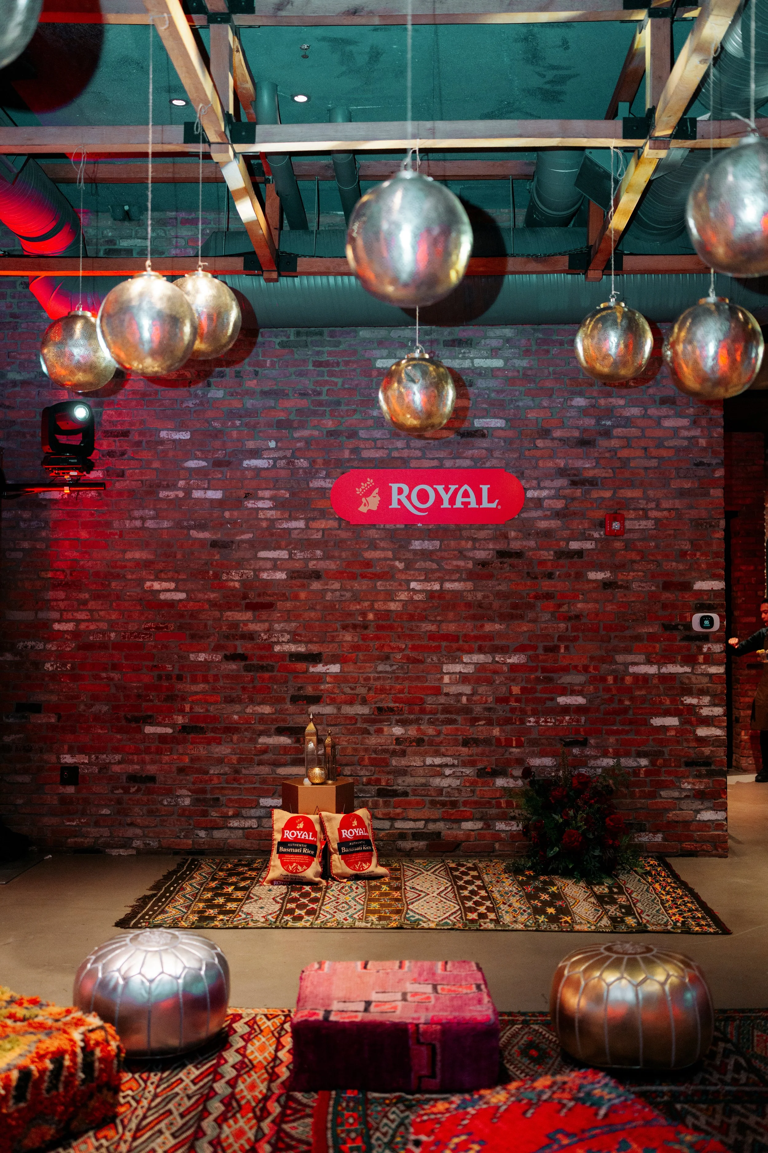 Decorative indoor display with hanging metallic spheres, rugs, and a brick wall with a red sign reading 'Royal'. There are decorative pillows, lanterns, and plants on the floor.