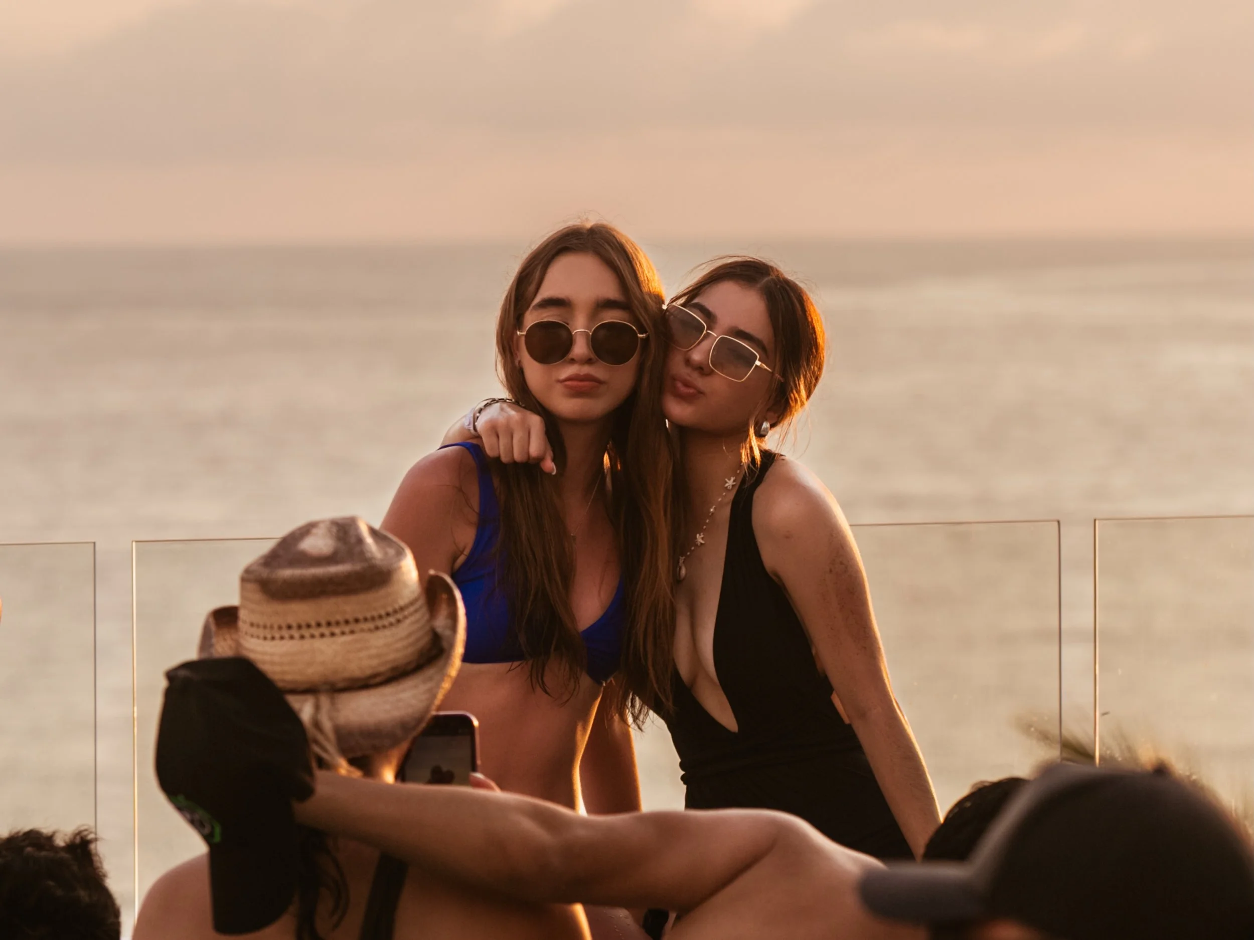 Dos mujeres posando en un beach club durante el atardecer, una con gafas redondas y otra con gafas cuadradas, ambas en bikini, con el mar de fondo.