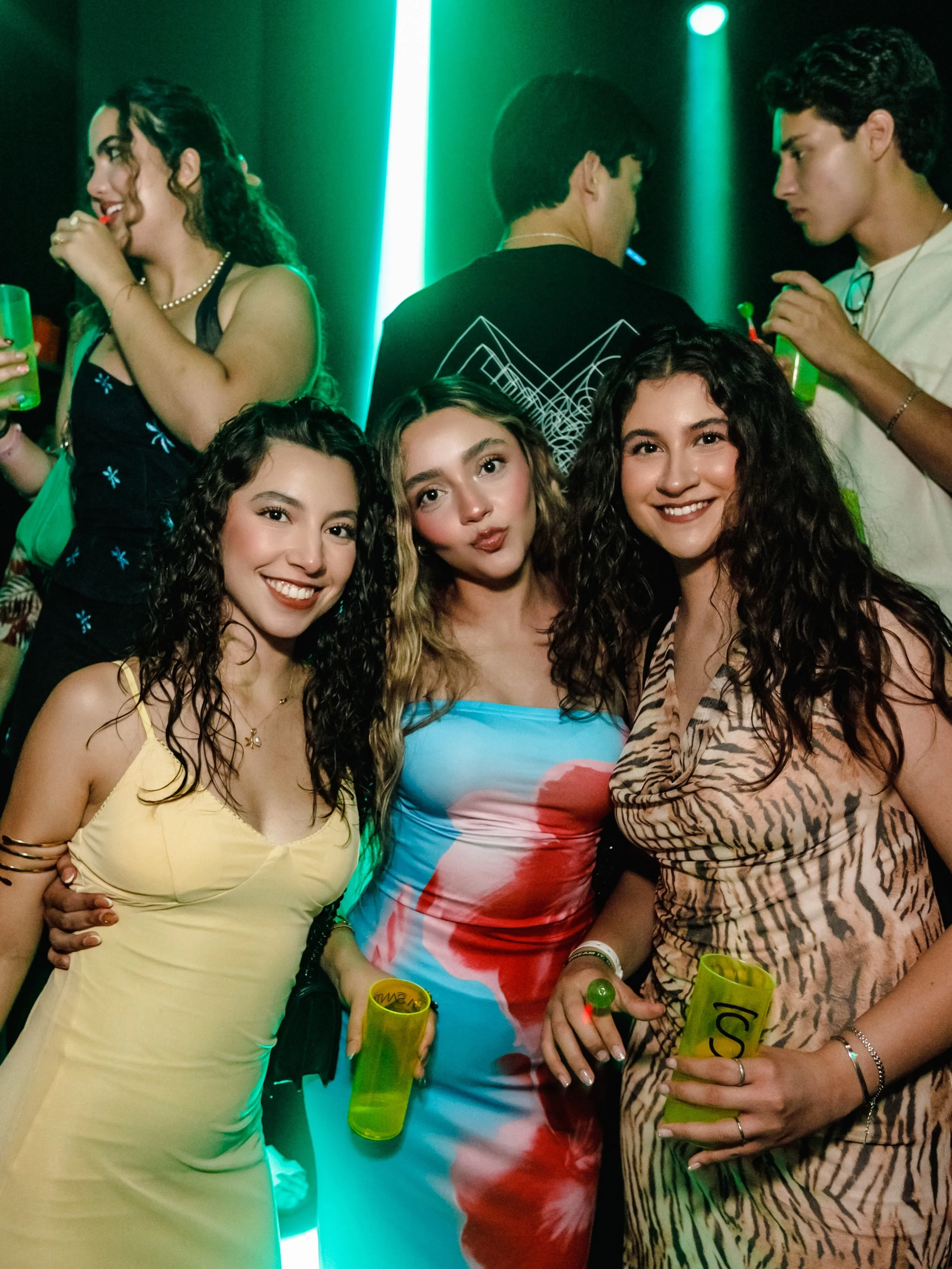Tres mujeres jóvenes sonriendo y sosteniendo vasos en una fiesta, con fondo de luces de neón y personas bailando.