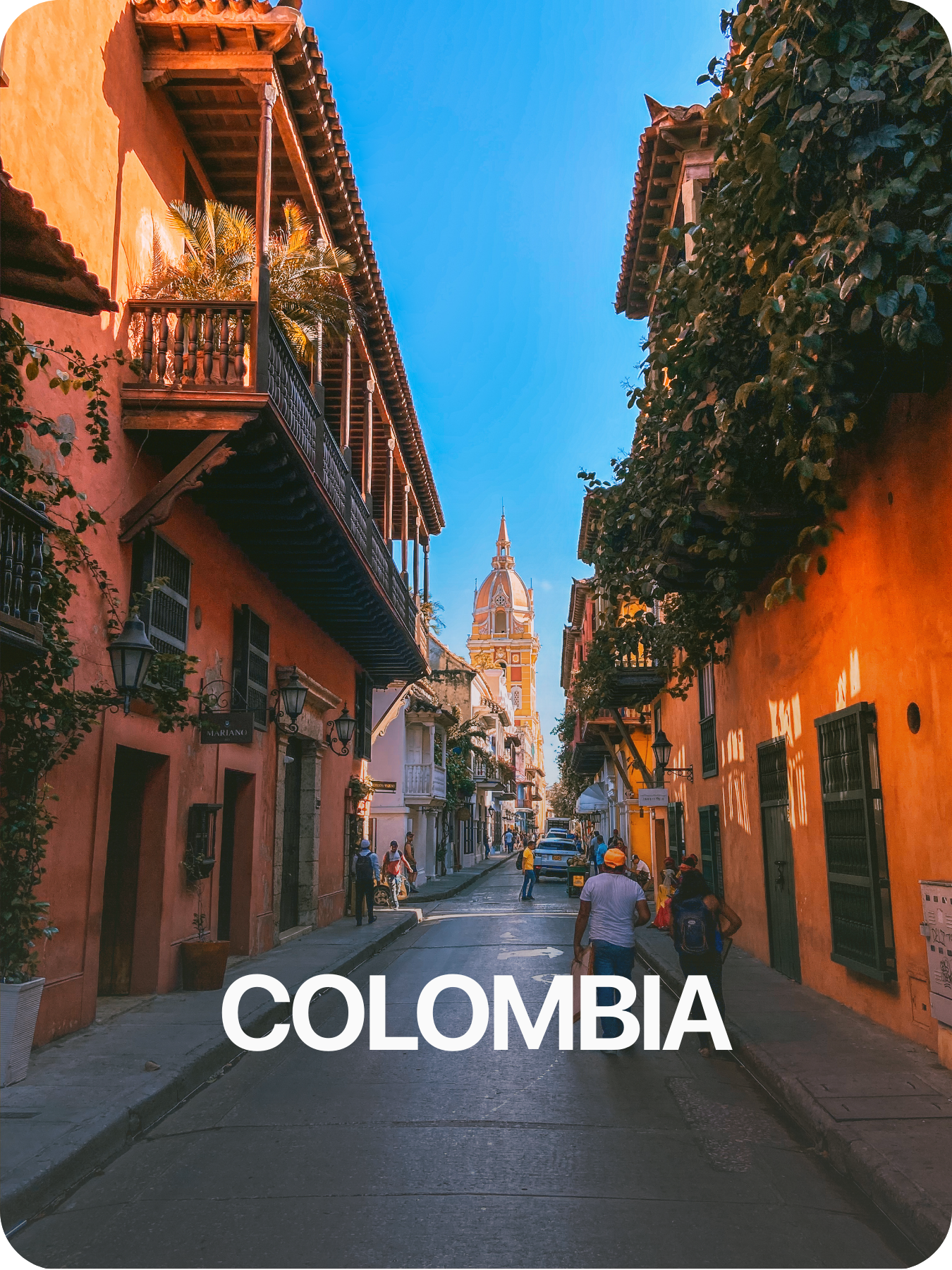  calle empedrada con edificios de colores cálidos y balcones de madera, en el fondo una iglesia con torre y pináculo, personas caminando y autos estacionados, clima soleado y cielo azul