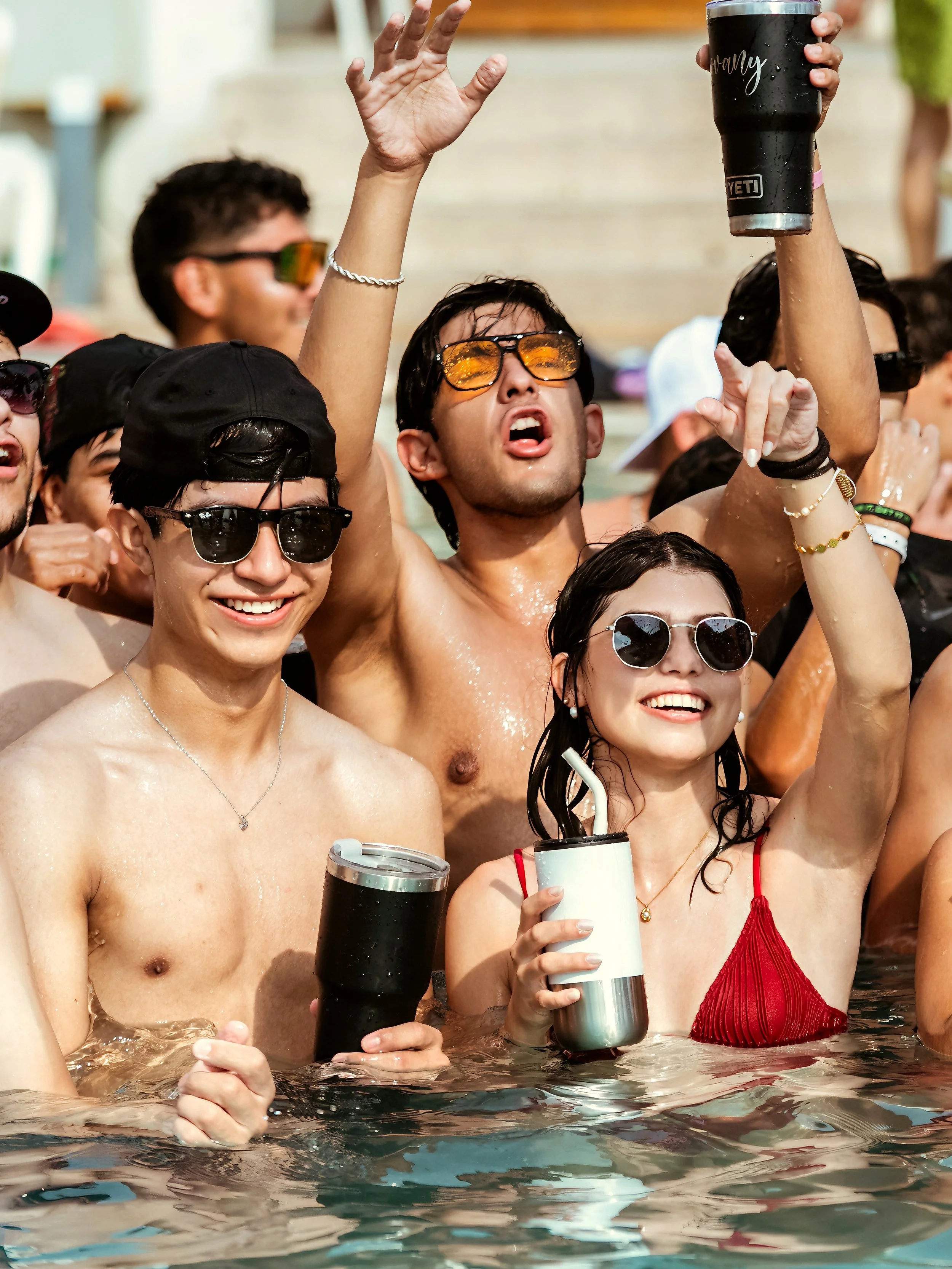 Grupo de jóvenes en una piscina celebrando, algunos con gafas de sol y cervezas en manos.