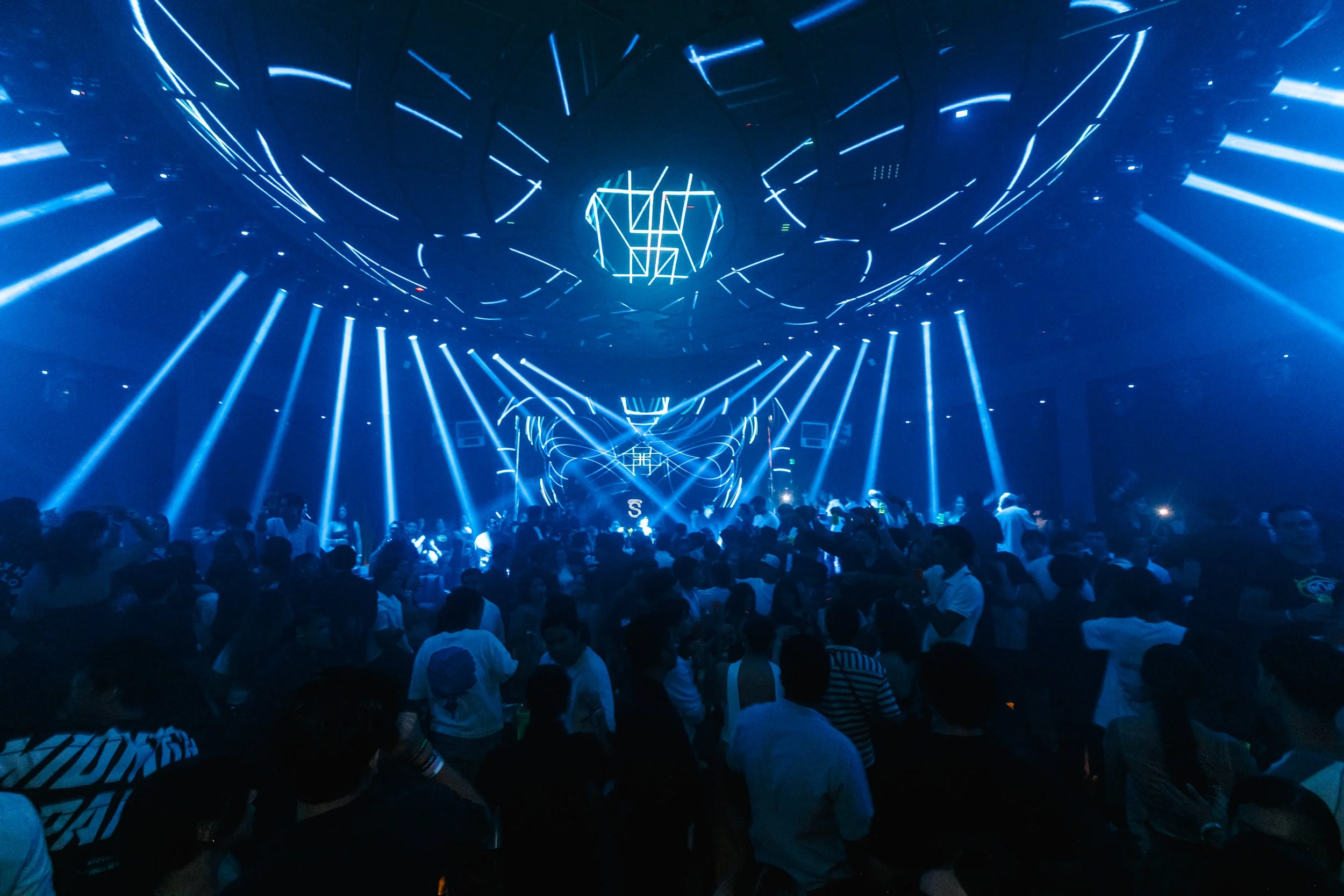 Una multitud en un club nocturno con luces azules y un escenario con diseño luminoso.
