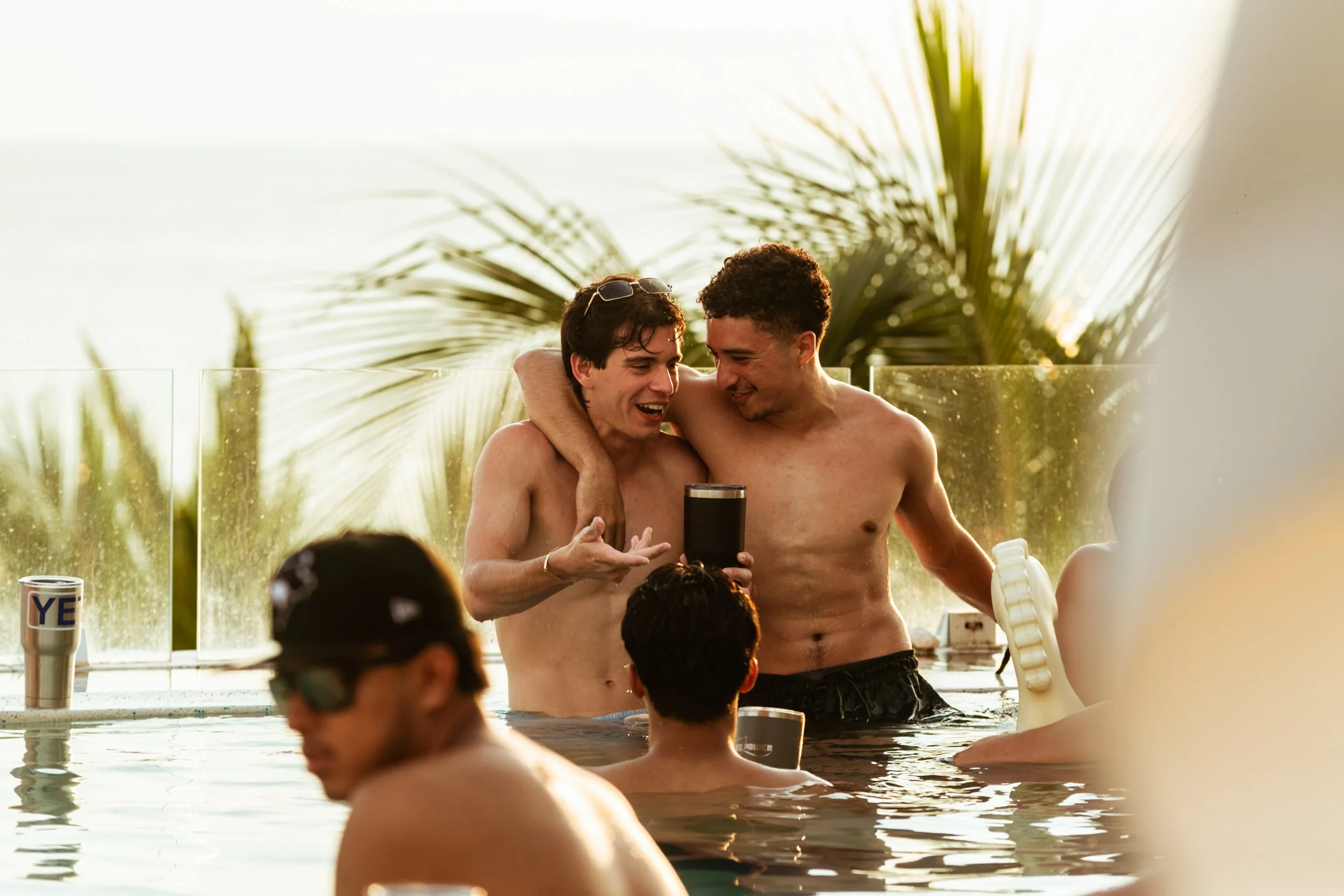 Grupo de jóvenes disfrutando en una piscina, algunos con gafas de natación, celebrando o compartiendo con bebidas, en un ambiente soleado con palmeras de fondo.