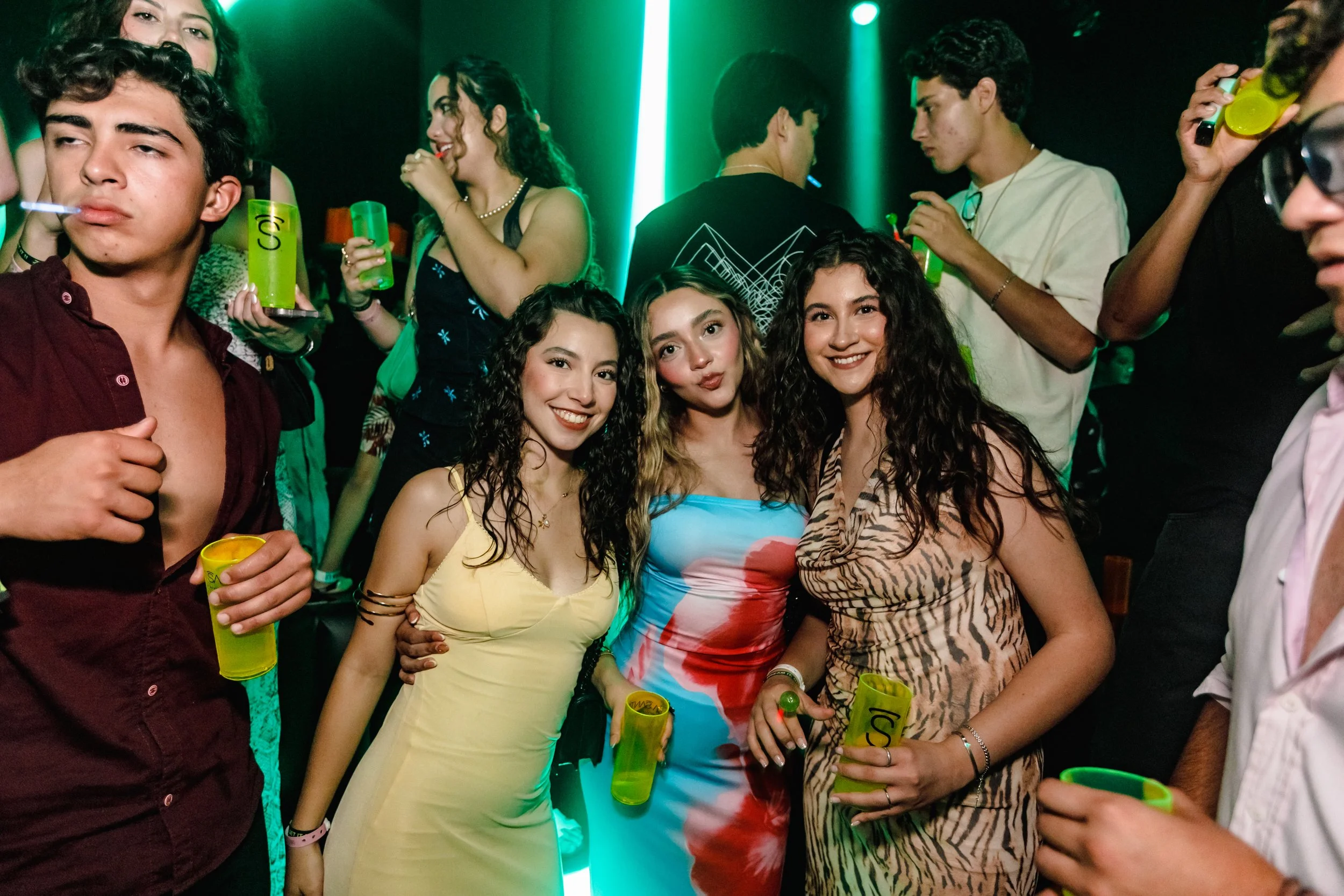 Grupo de jóvenes en una fiesta, sonriendo y sosteniendo vasos con bebidas, con luces verdes de fondo.