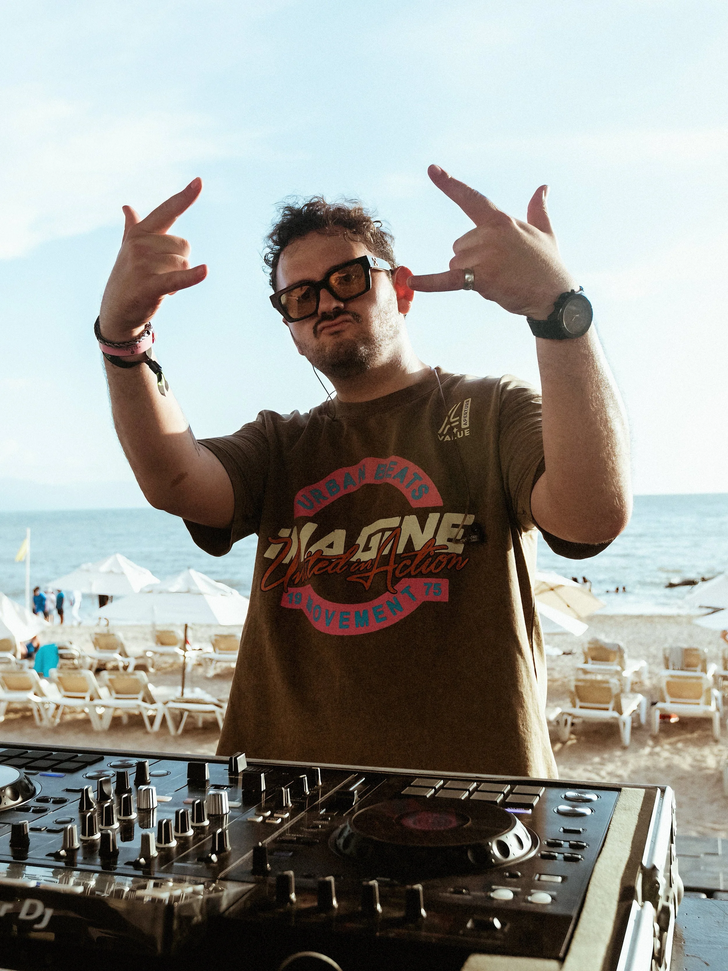 Persona frente a equipo de DJ en la playa haciendo señal de paz con las manos, fondo de arena y sillas con sombrillas cerca del mar.