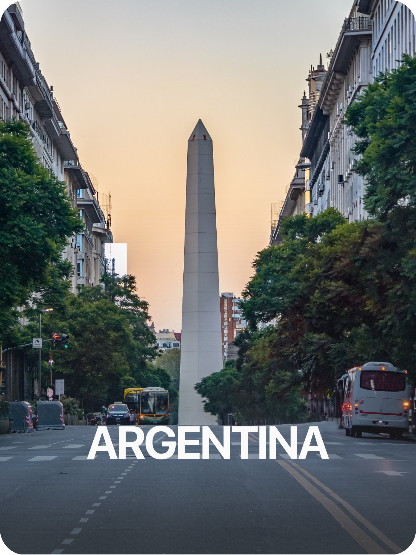 Vista de la avenida con árboles y transporte público, en el fondo el Obelisco de Buenos Aires, en Argentina, durante el atardecer.