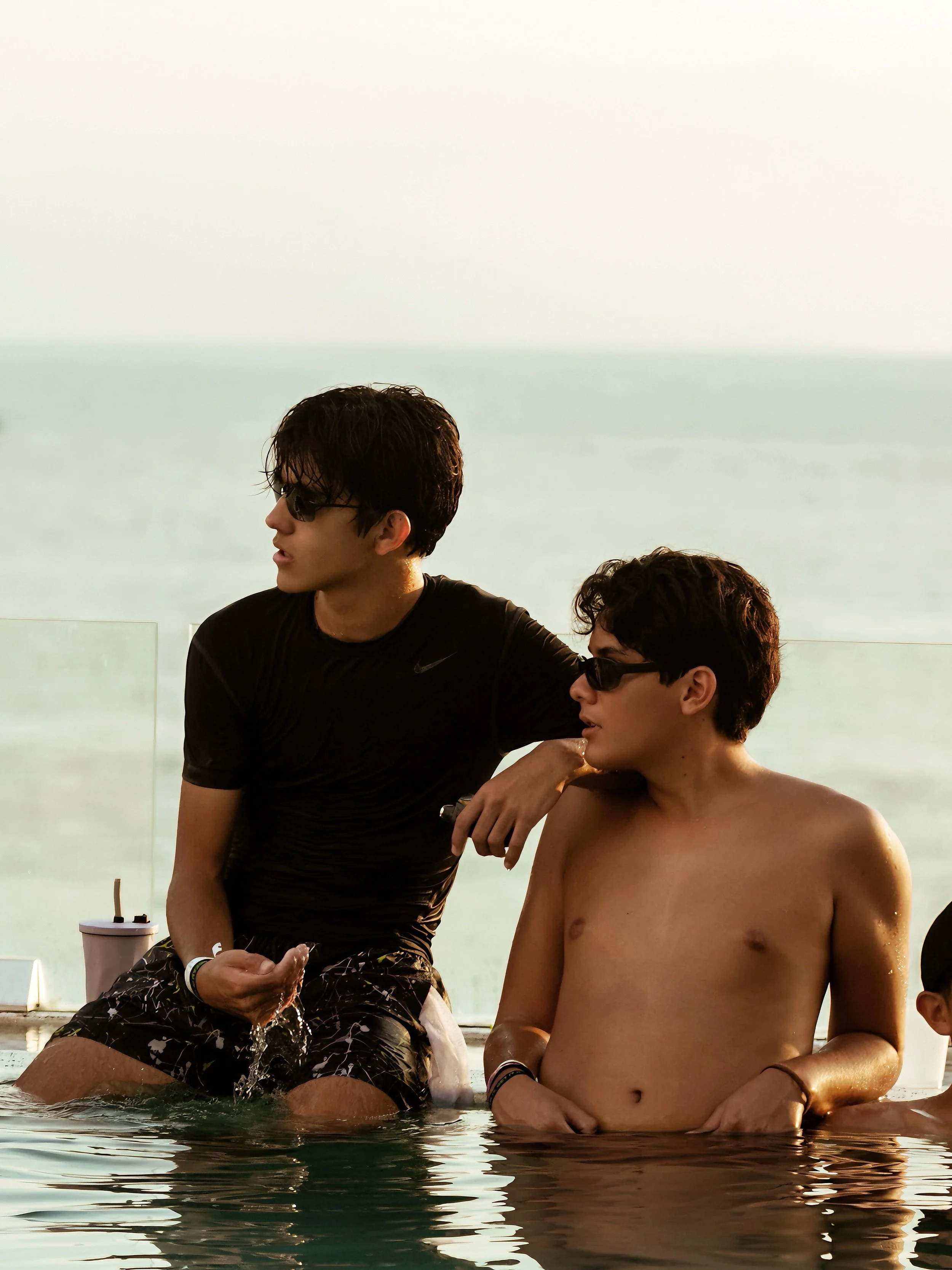 Dos jóvenes en una piscina, uno de ellos joven sin camiseta y otro con camiseta negra y gafas de sol, con vista al mar de fondo.