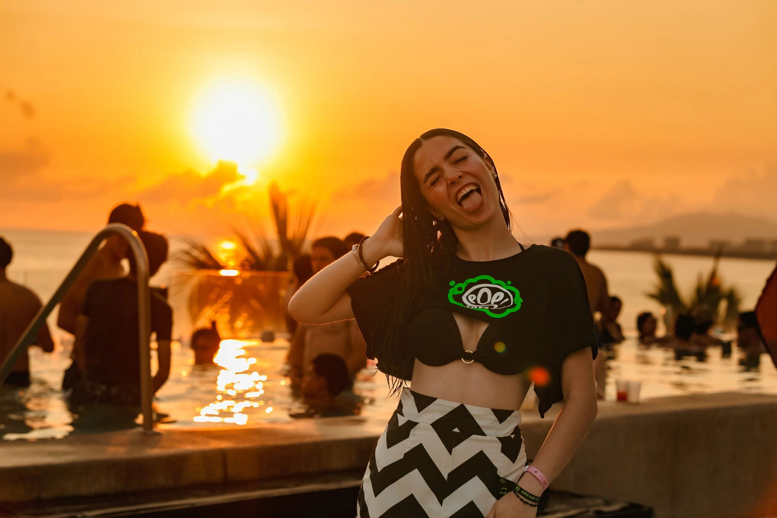 Joven mujer en una fiesta en la piscina disfrutando del atardecer, con amigos al fondo y expresión feliz.
