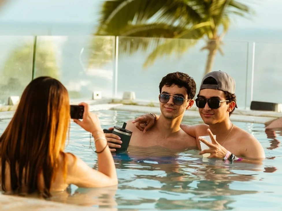 Dos jóvenes en la piscina posando para una foto con una mujer, con fondo de palmera y vista al mar.