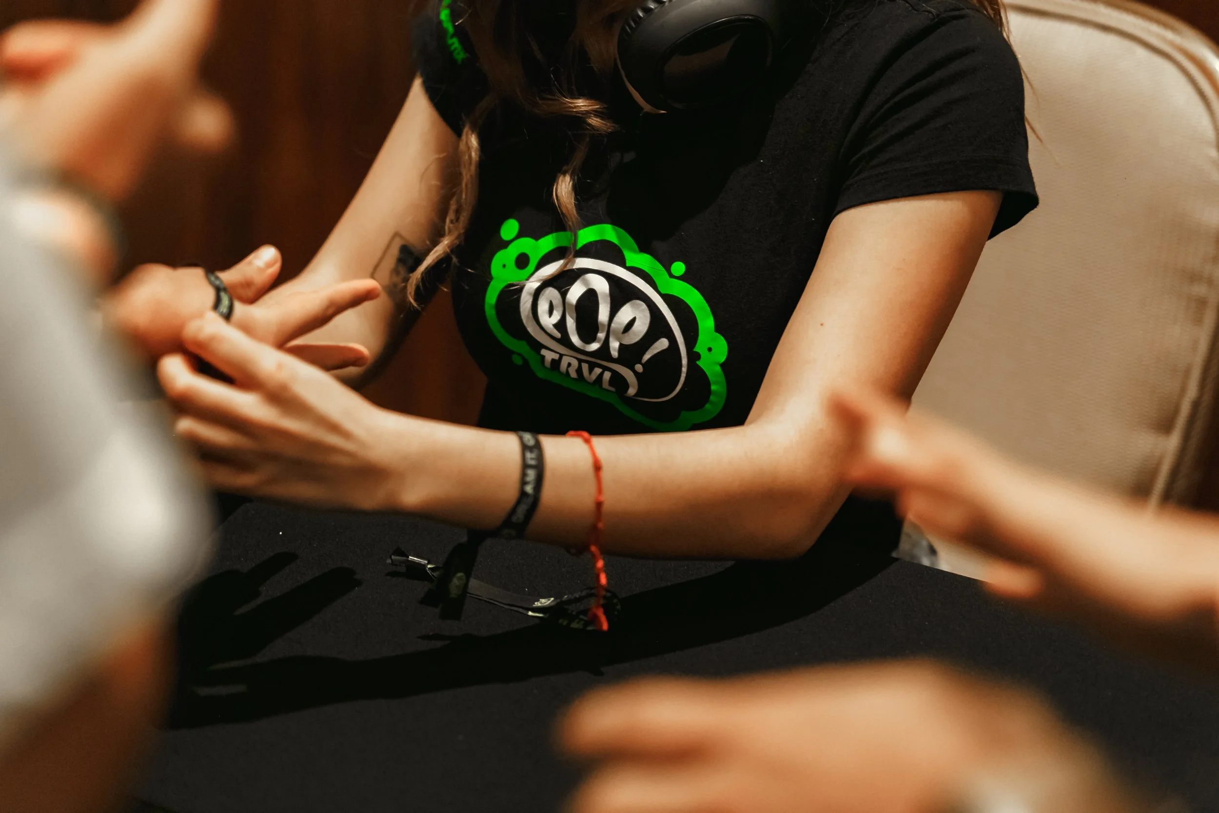 Persona con camiseta negra que tiene un logotipo en verde y blanco, sentada en una mesa con un fondo oscuro, rodeada de otras personas cuyos brazos y manos están en primer plano.