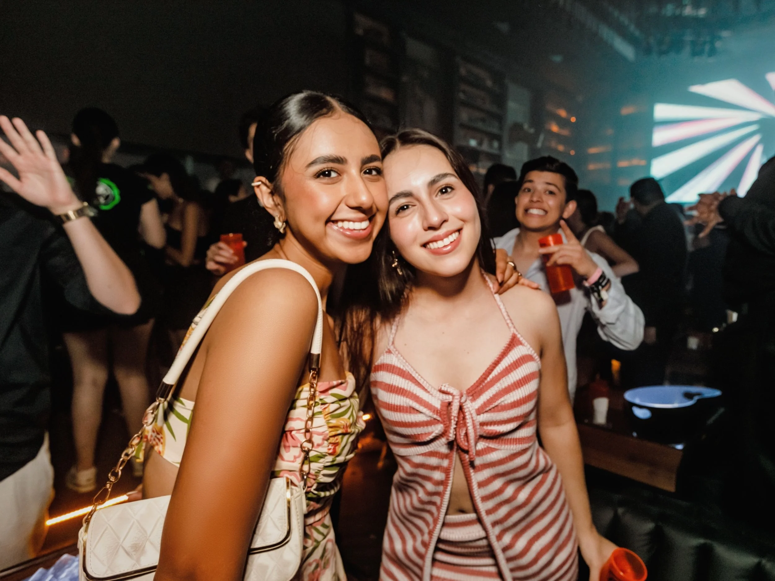 Dos mujeres sonriendo en un ambiente de fiesta, rodeadas de personas bailando y luces de neón.