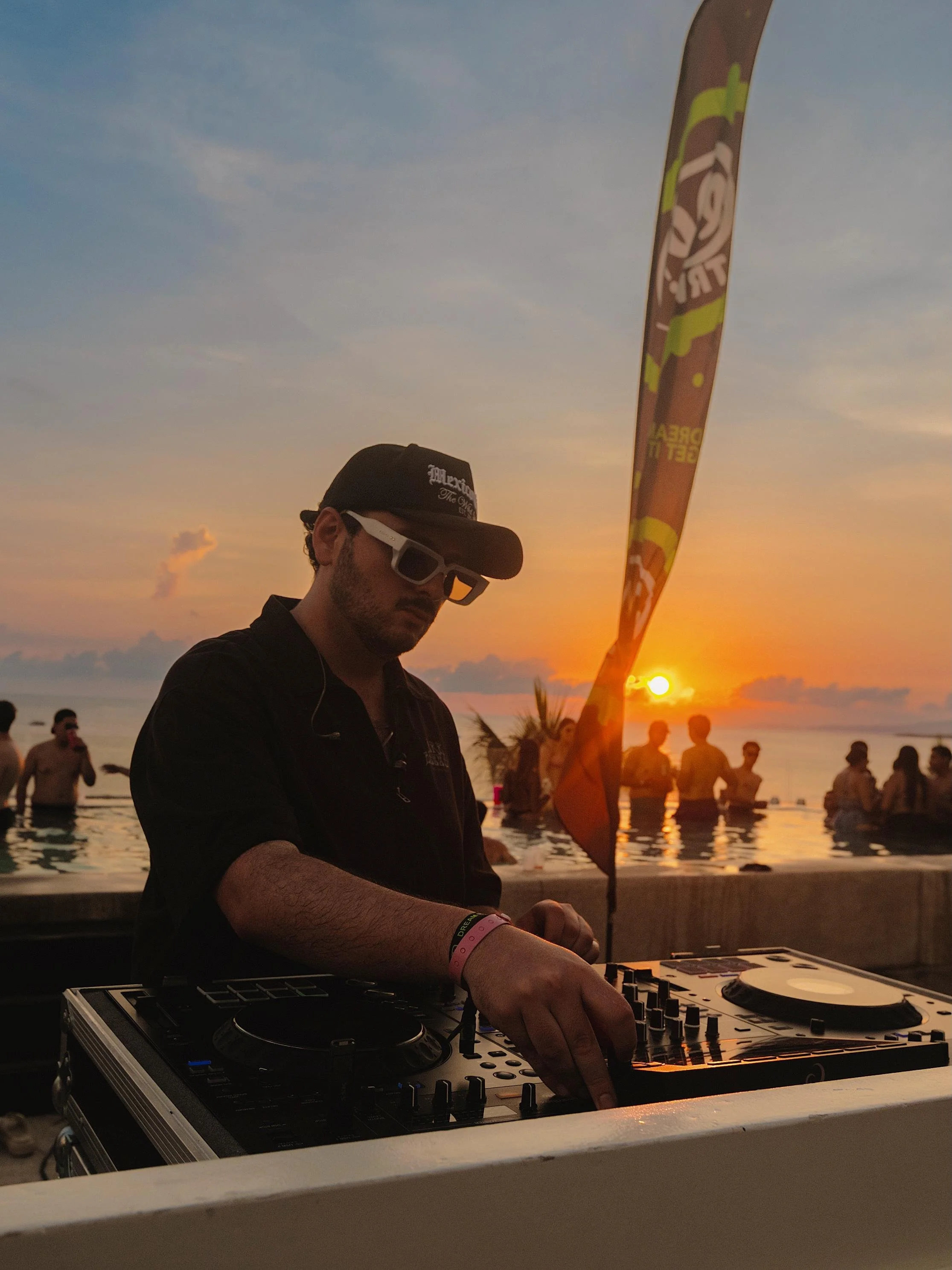 Un DJ tocando música en una fiesta junto a la playa al atardecer, con personas en el fondo disfrutando del ambiente.