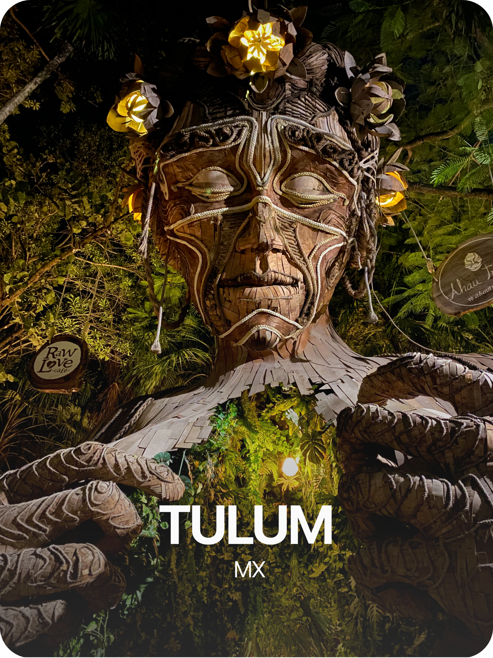 Escultura de una figura humana indígena tallada en madera con detalles de cuerda y decoraciones florales, rodeada de vegetación y luces, en Tulum, México.
