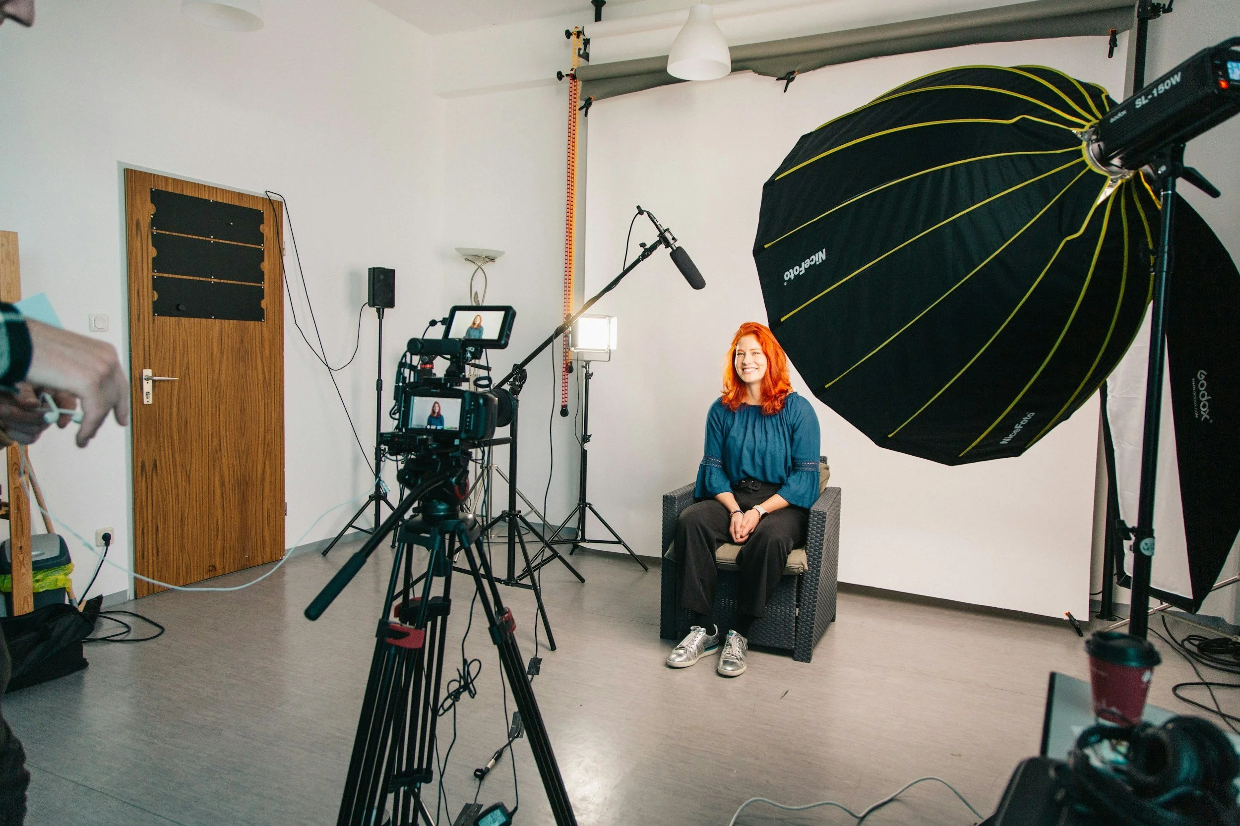 A woman with red hair is sitting on a chair in front of a white wall in a photography studio. She is smiling and wearing a blue blouse and black pants. There is professional lighting and camera equipment around her, including softboxes, a camera on a tripod, a large reflector, and other accessories.
