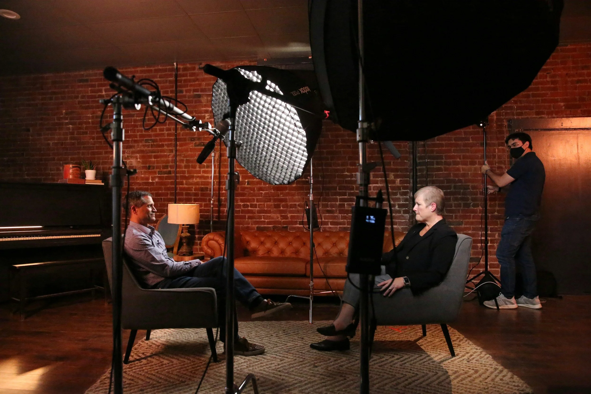 Two people seated in chairs facing each other in a recording studio with a brick wall background, while a crew member adjusts lighting equipment.