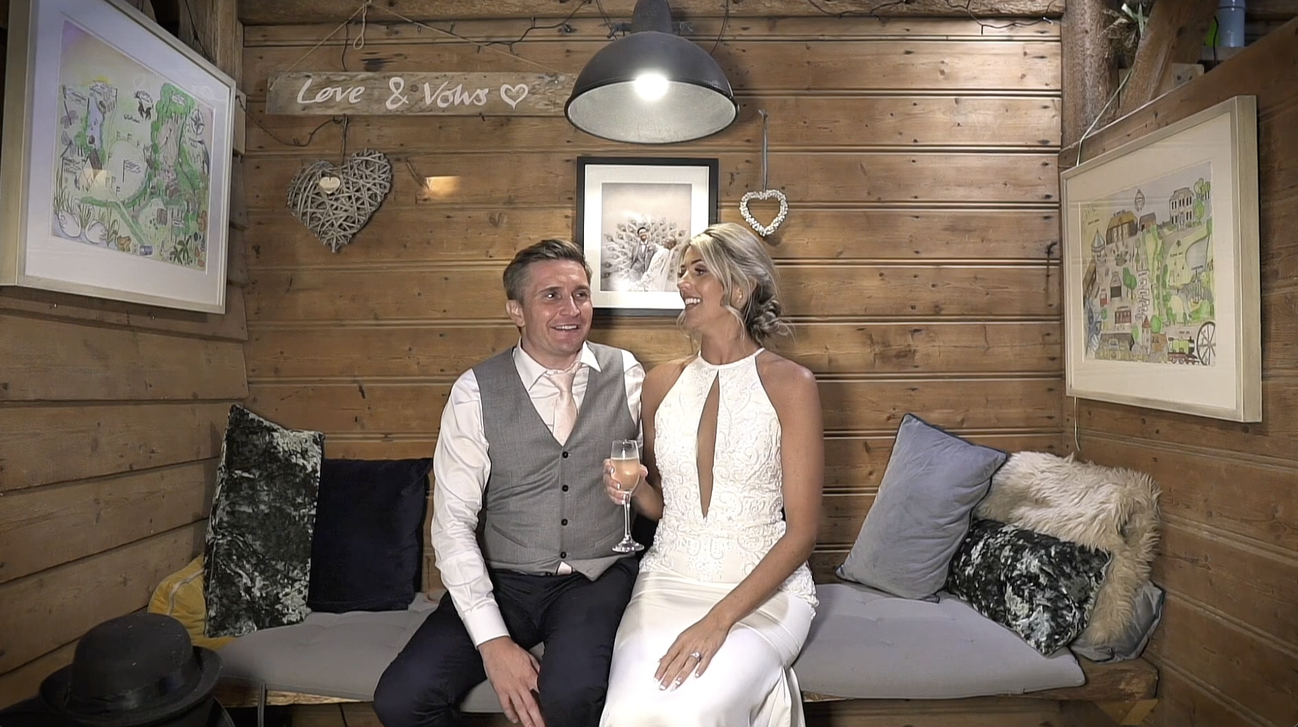 A couple sitting on a bench in a rustic wooden room, smiling and looking at each other. The man is wearing a gray vest, white shirt, and black pants, holding a glass of champagne. The woman is in a white dress with lace details. The room is decorated with framed artwork, heart-shaped ornaments, and a sign that says 'Love & Vows'.