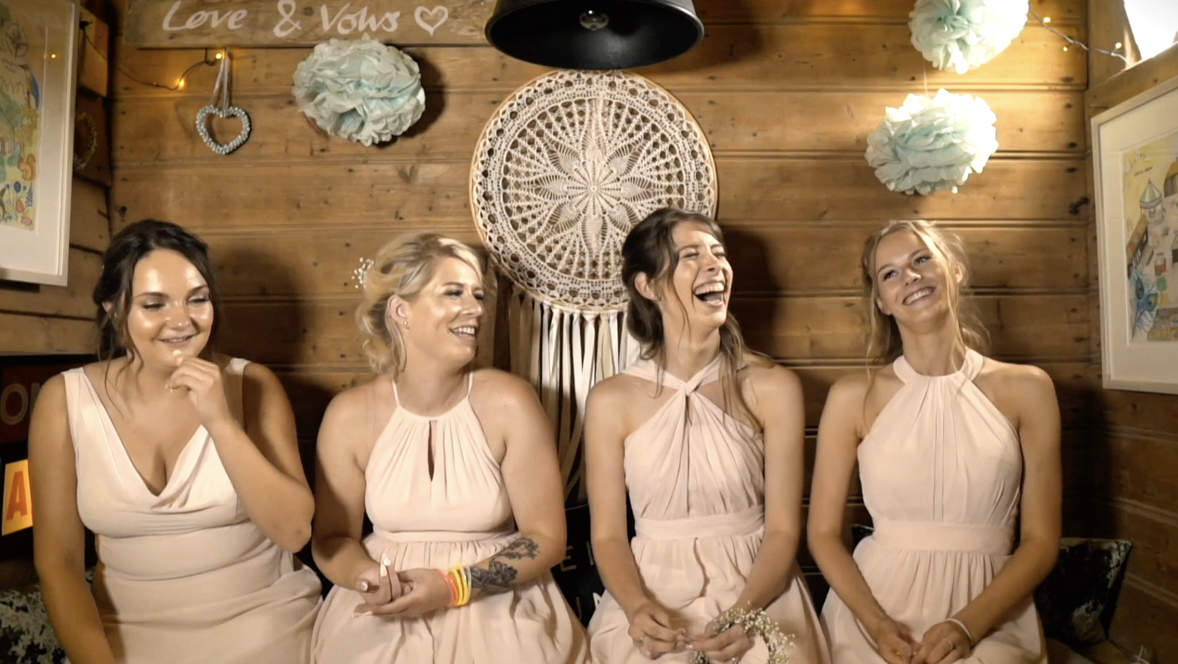 Four women wearing matching beige dresses sit in front of a wooden wall decorated with paper pom-poms, a dreamcatcher, and framed art, smiling and laughing during what appears to be a celebration.