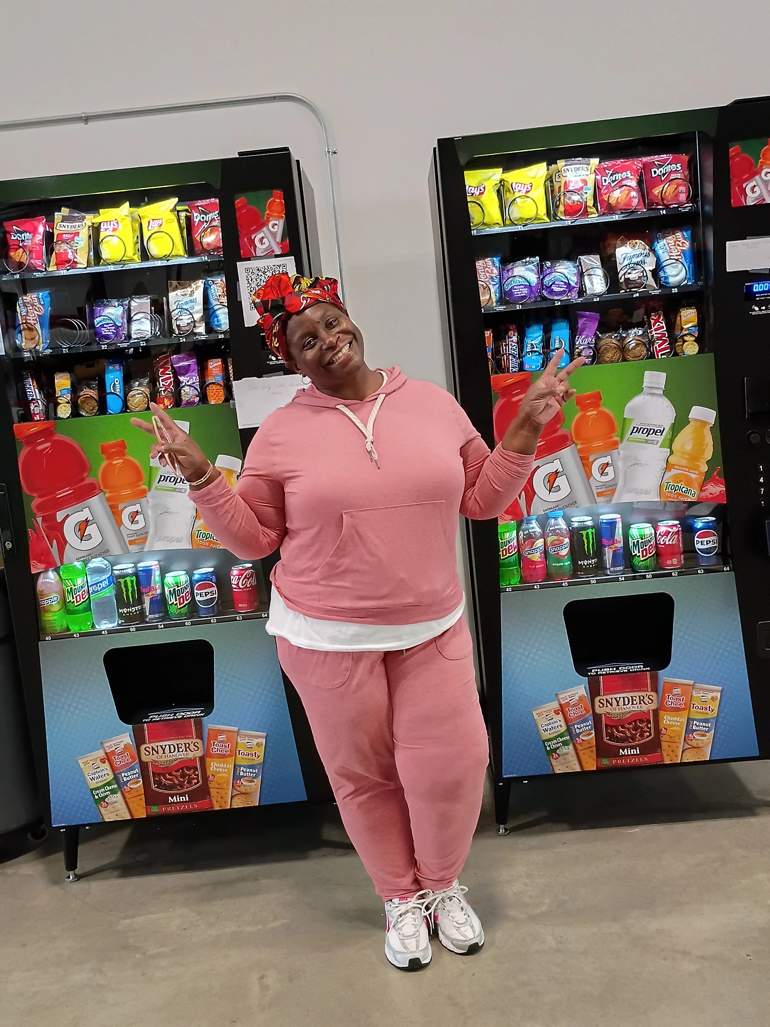 A woman dressed in pink tracksuit and white sneakers, smiling and posing with a peace sign in front of vending machines filled with snacks and beverages.