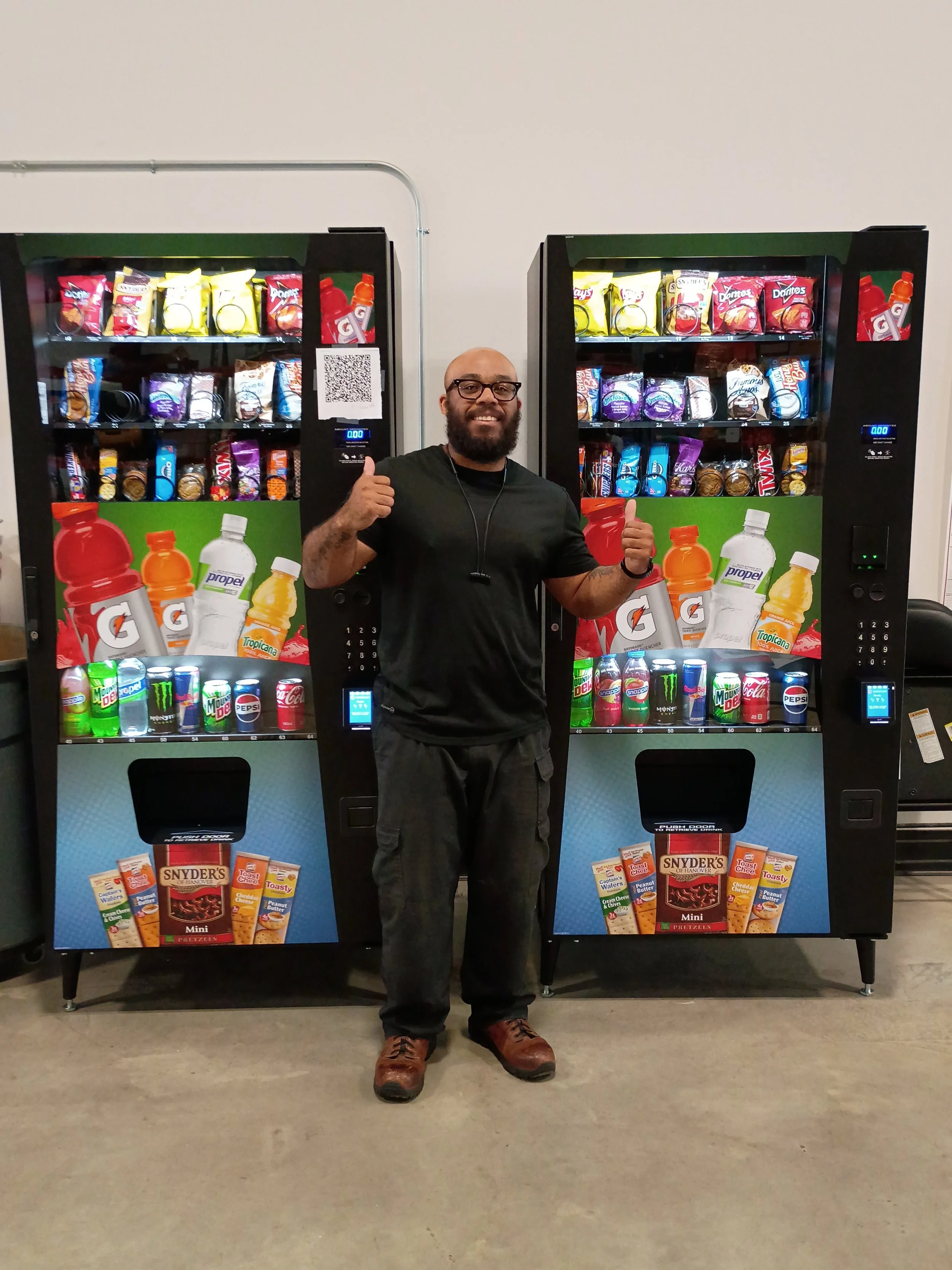 Man with glasses and beard smiling and giving a thumbs up while standing in front of two snack and soda vending machines.