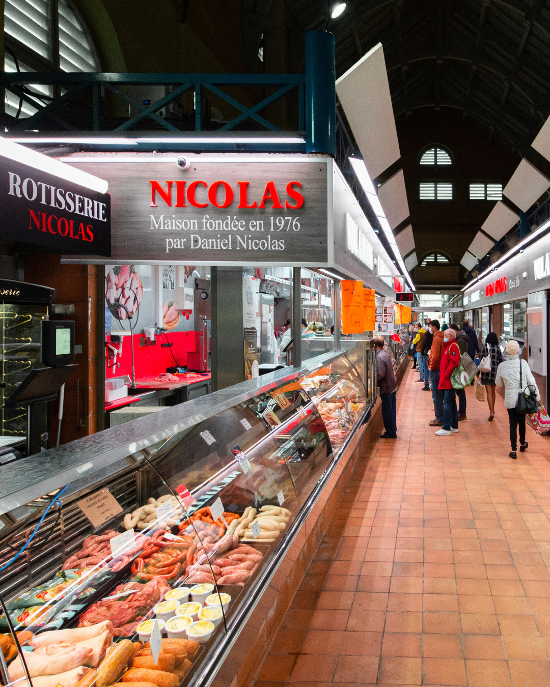 Intérieur d'un marché, rayon de vente de produits de boucherie avec des clients en file d'attente. Signage indiquant la maison Nicolas fondée en 1976 par Daniel Nicolas.