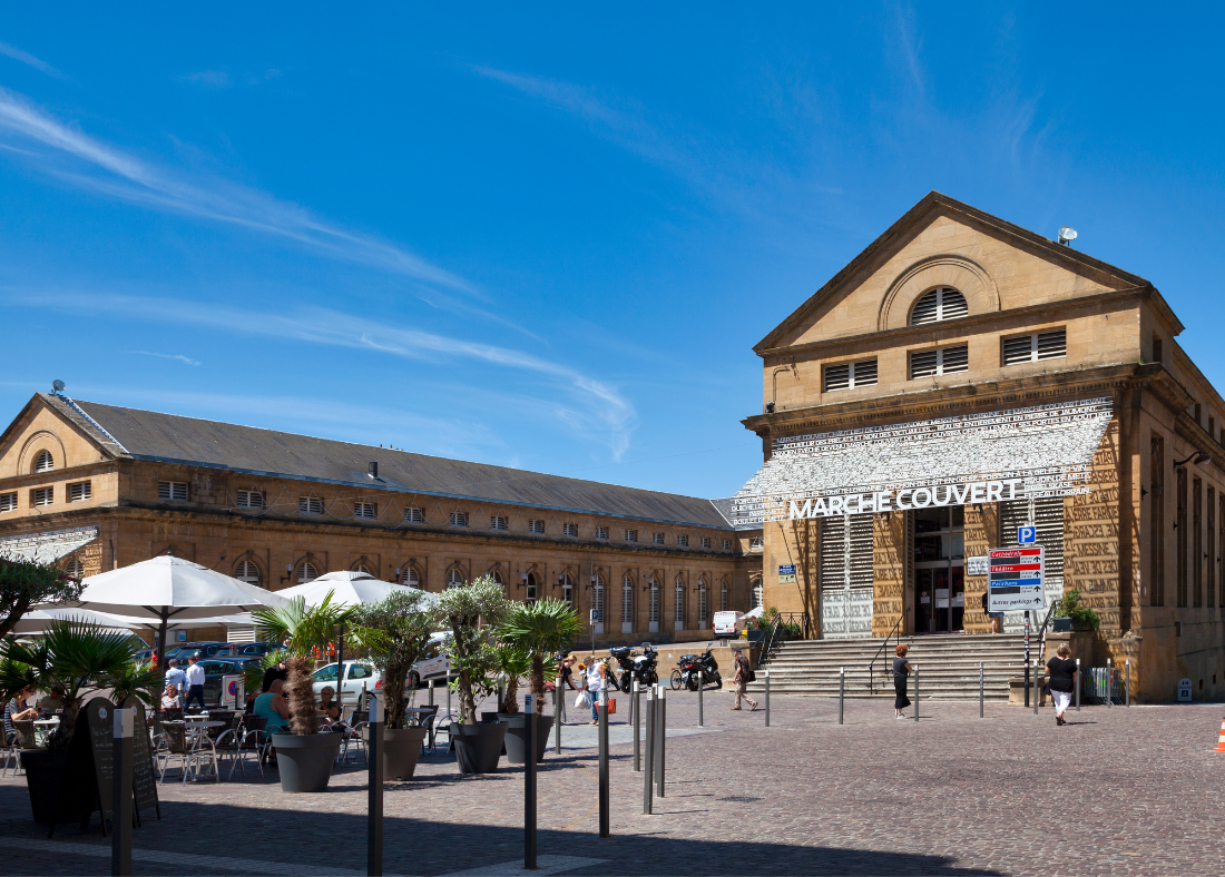 Bâtiment en pierre avec inscription "MARHE-COUVERT", devant lequel des gens circulent, sous un ciel bleu.