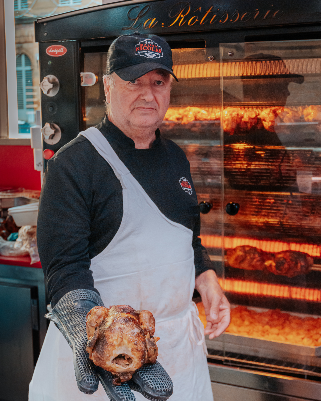 Homme portant un tablier blanc et un chapeau noir, tenant un poulet croustillant devant une rôtissoire