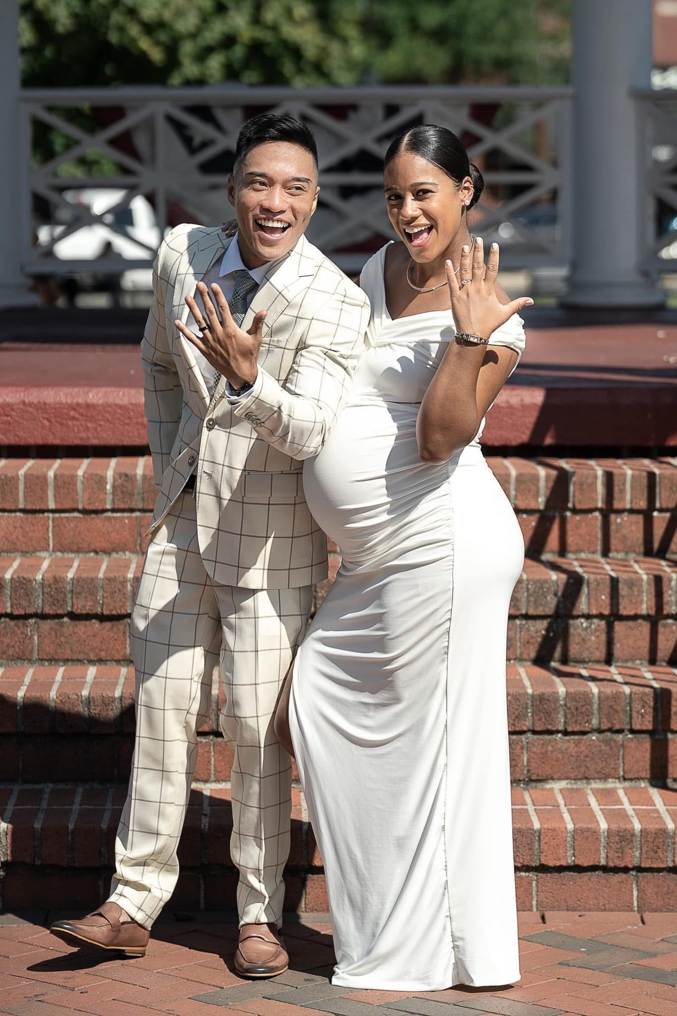 A joyful couple, man in a beige checked suit and woman in a white dress, celebrating outdoors on stairs, showing off rings on their hands.