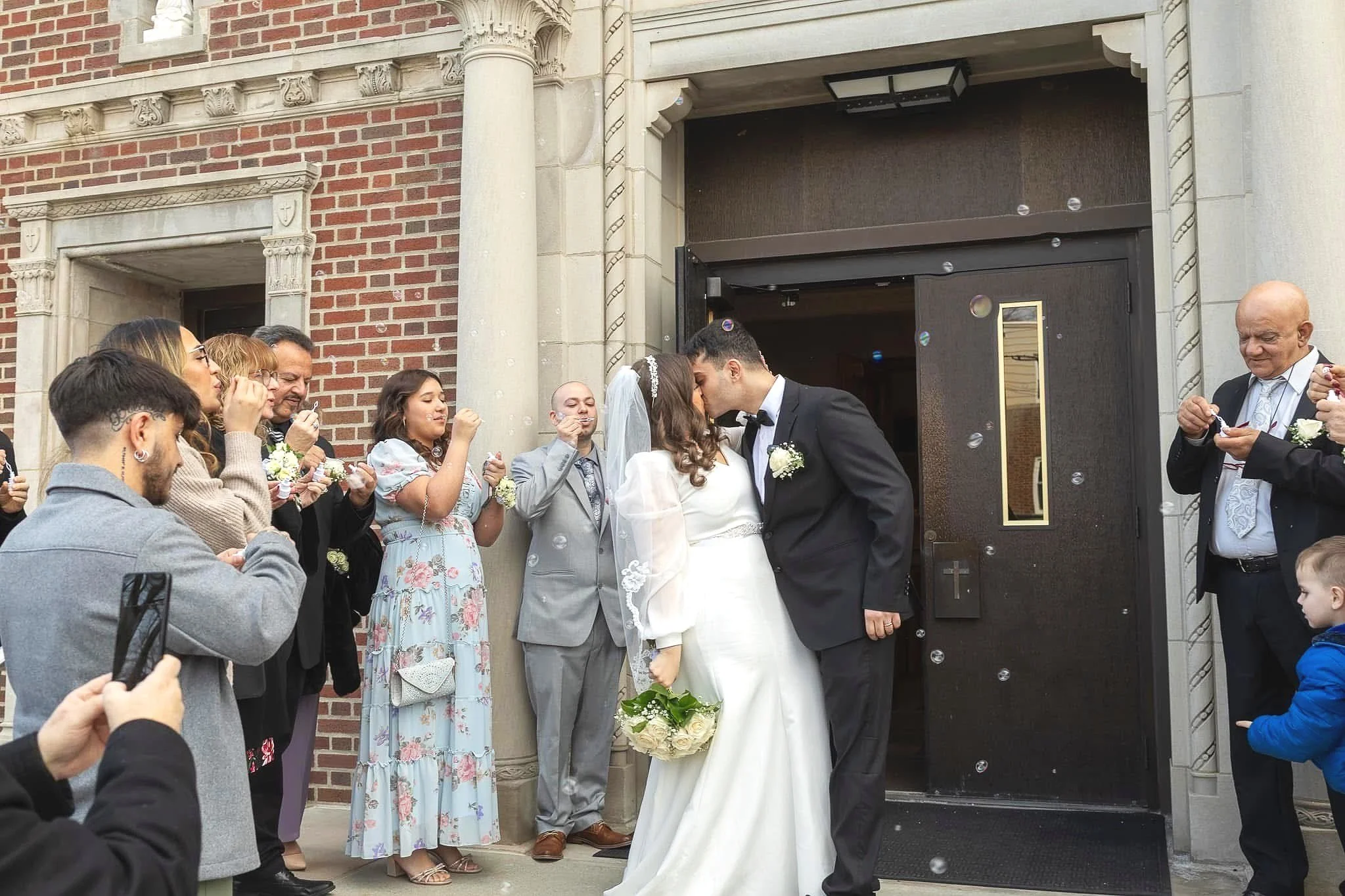 A newlywed couple sharing a kiss outside a church entrance surrounded by family and friends celebrating their wedding.