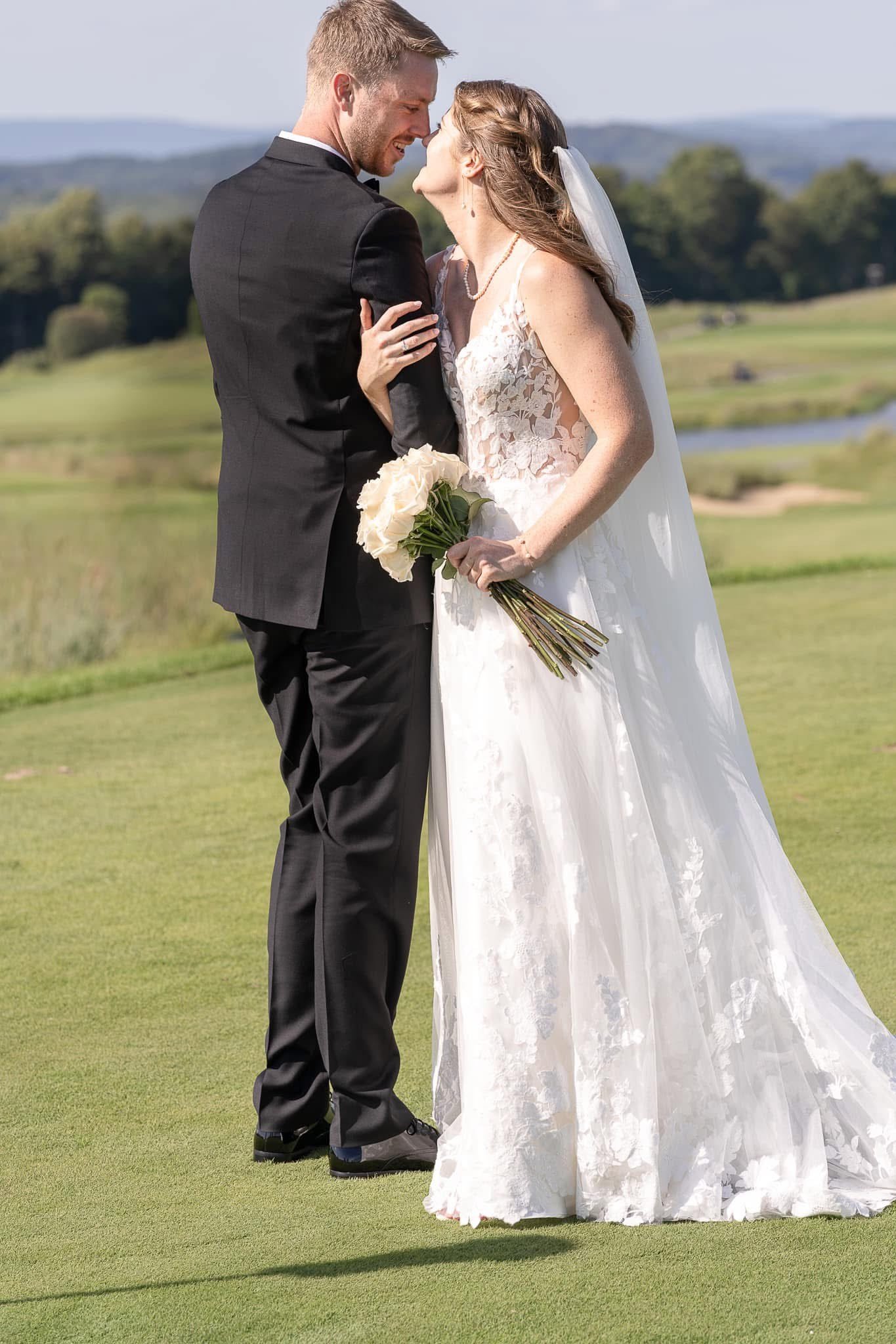A bride and groom embrace outdoors on a golf course, with the bride holding a white bouquet, both smiling and about to kiss.