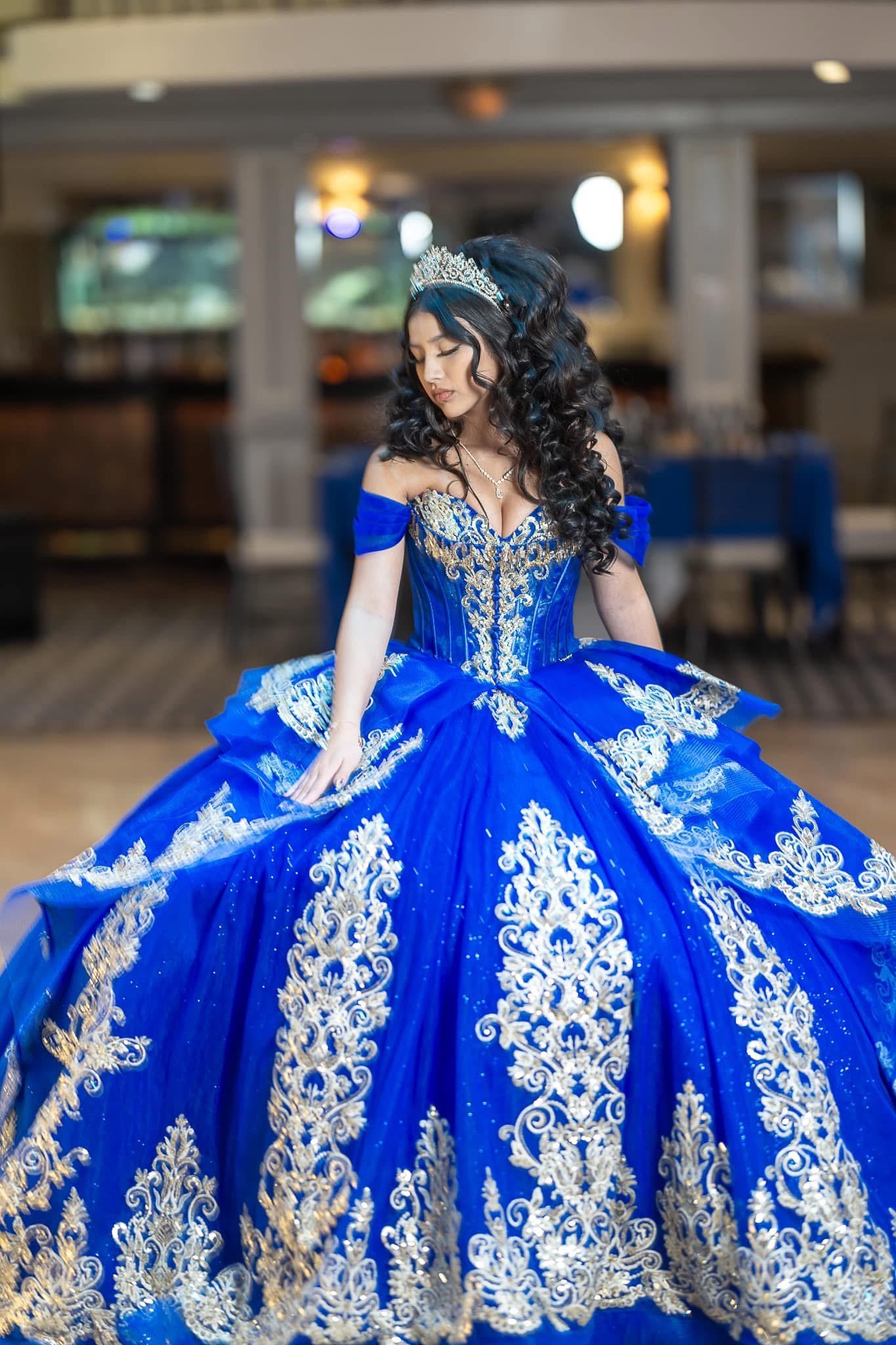 Young woman in a royal blue gown with elaborate gold embroidery, wearing a tiara, standing indoors with a blurred background.