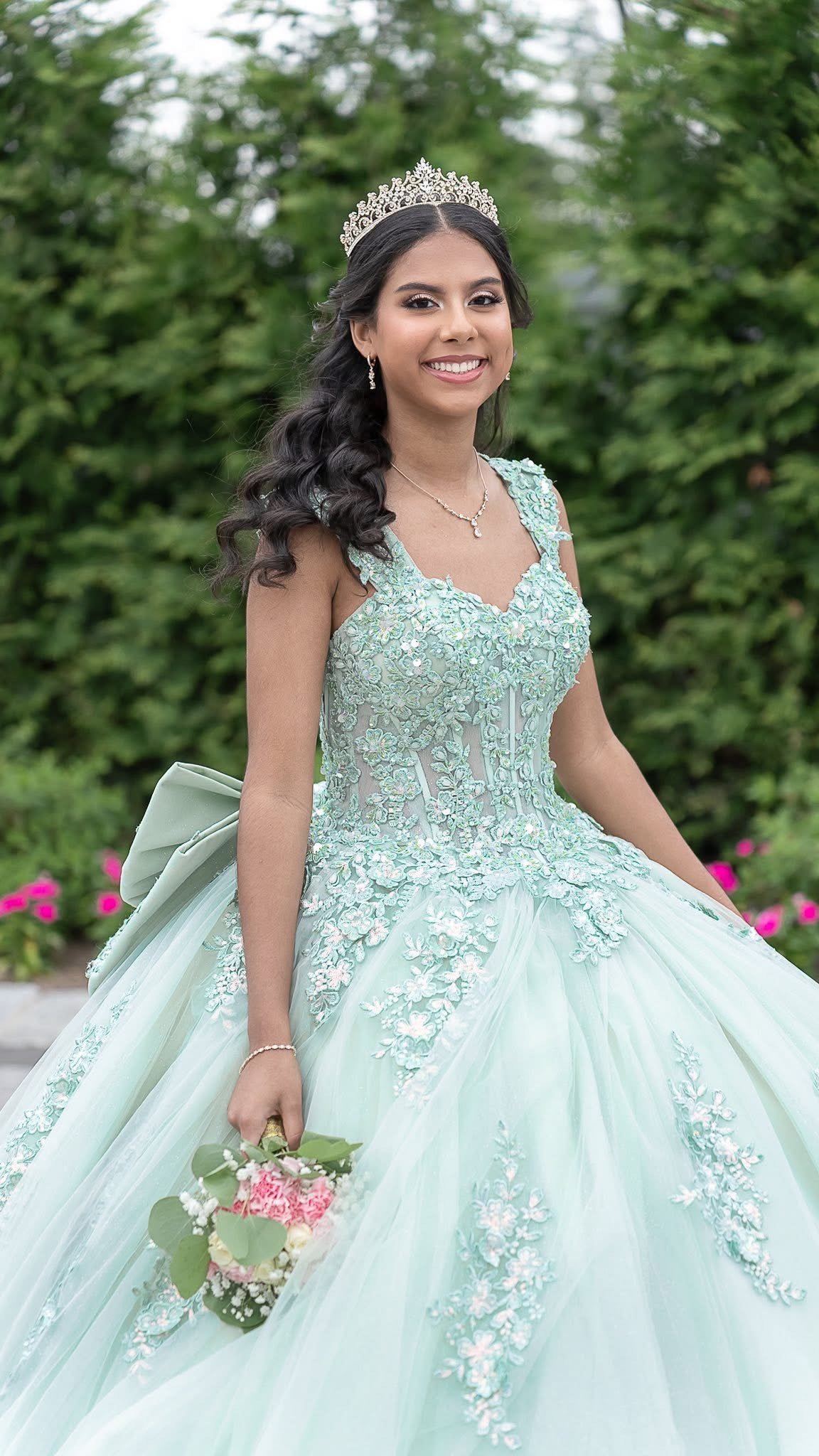 A young woman in an ornate, light turquoise ball gown with floral embroidery, wearing a tiara and holding a small bouquet of pink and white flowers, standing outdoors with green trees in the background.