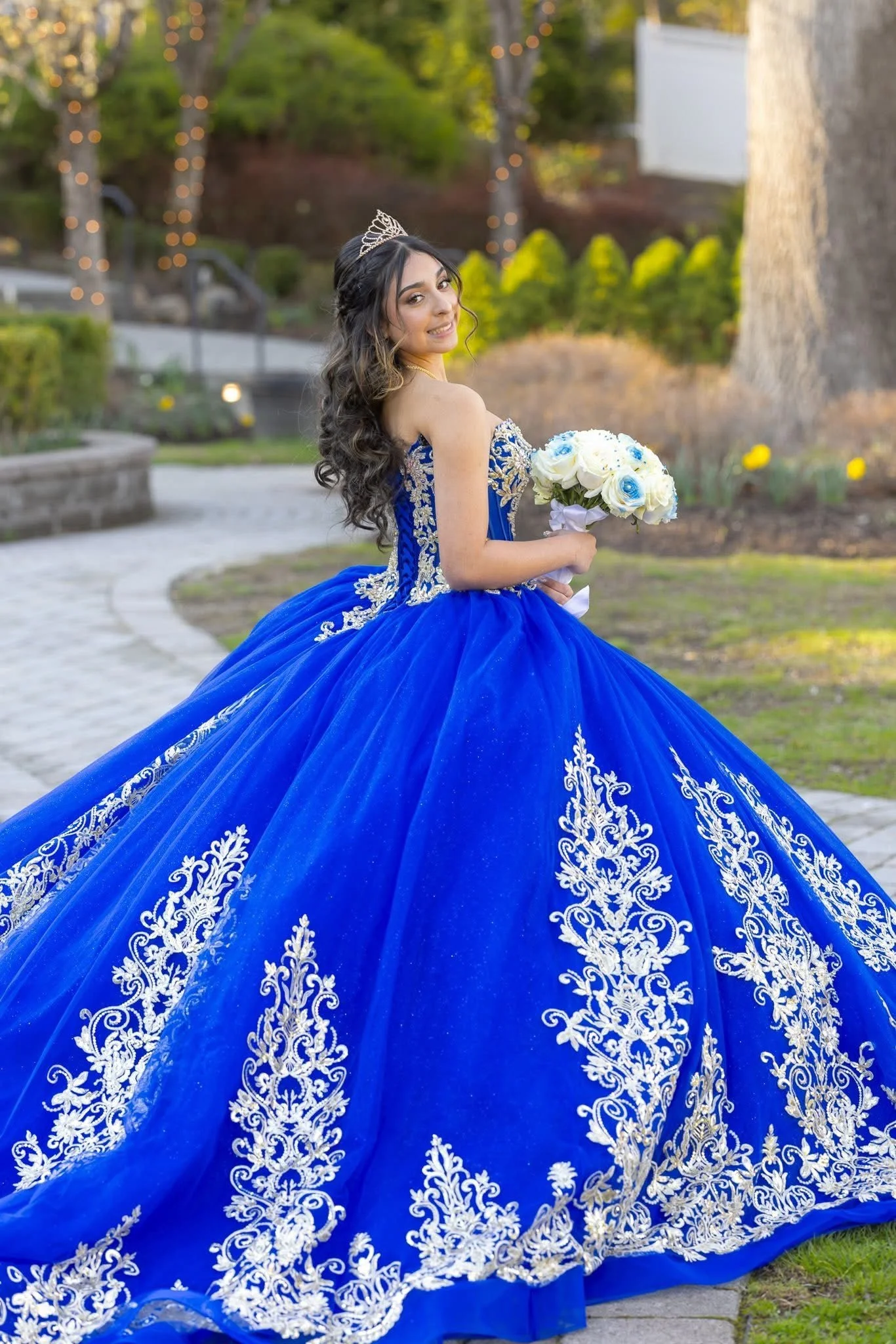 A young woman in a royal blue quinceañera gown with white embroidery and beadwork, holding a bouquet of white and blue flowers, outdoors in a landscaped garden with trees and yellow flowers.