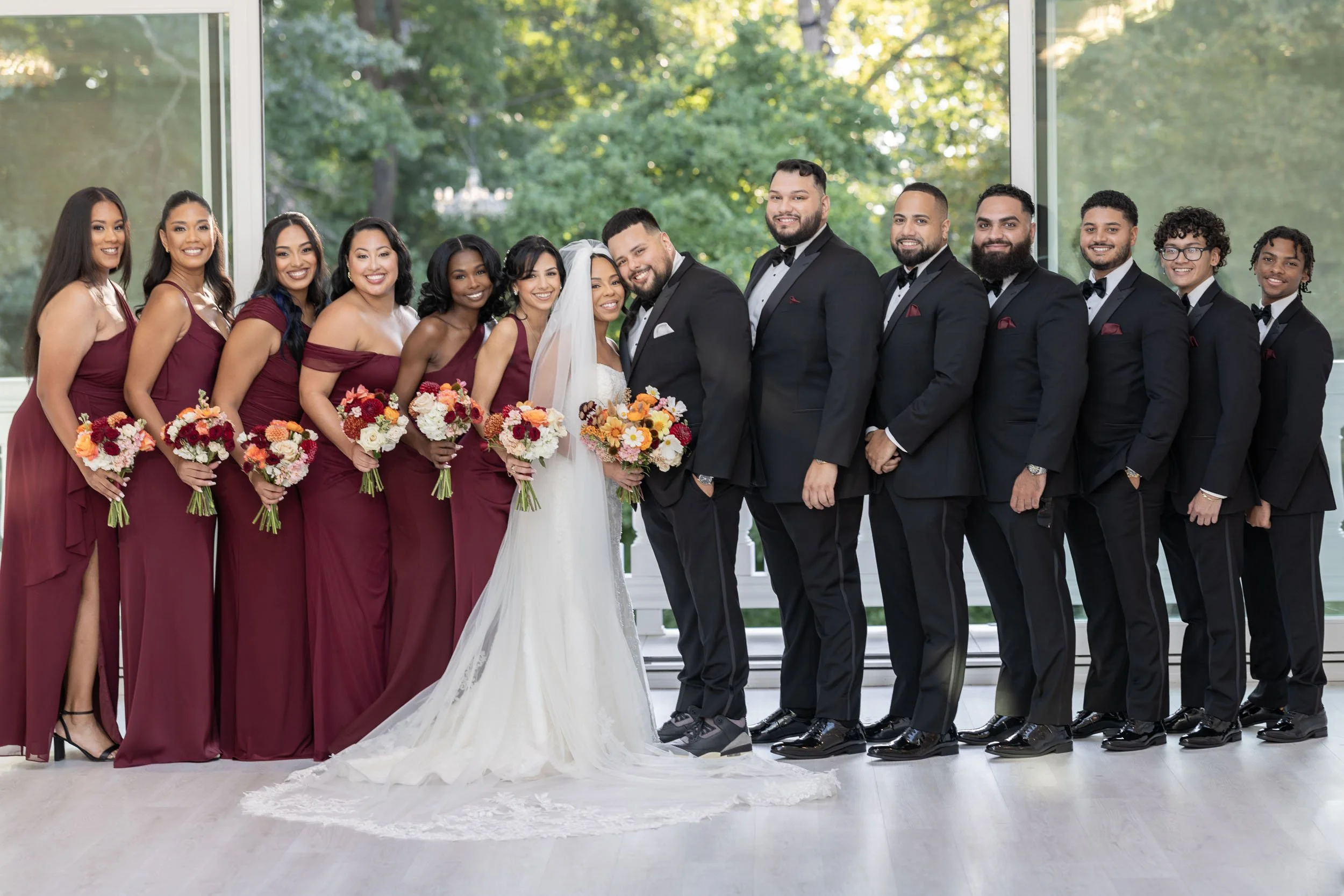 Group of wedding party members dressed in formal attire, with bridesmaids in burgundy dresses holding bouquets, groomsmen in black tuxedos, and a bride and groom in wedding attire, posing in front of large windows with a green outdoor backdrop.