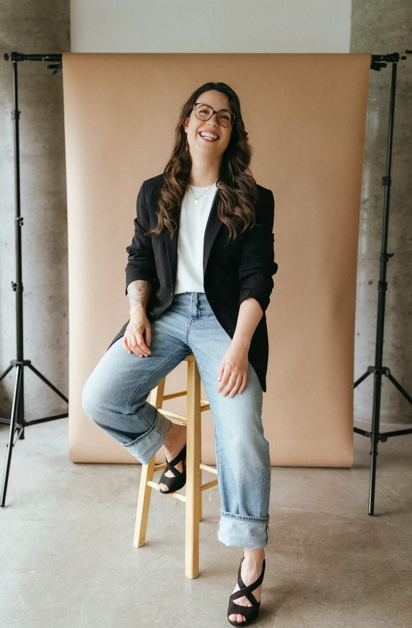 Une femme souriante assise sur un tabouret en bois, portant un blazer noir, un t-shirt blanc, un jean et des chaussures à talons noirs, devant un fond beige dans un studio.