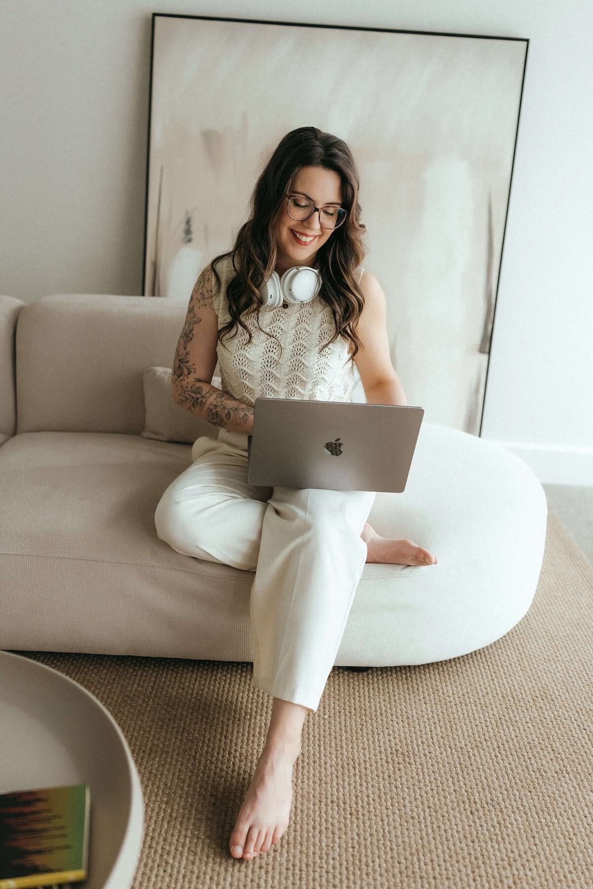 Jeune femme assise sur un canapé blanc avec un ordinateur portable, portant des lunettes, des écouteurs autour du cou, souriante, dans un environnement intérieur moderne et minimaliste.