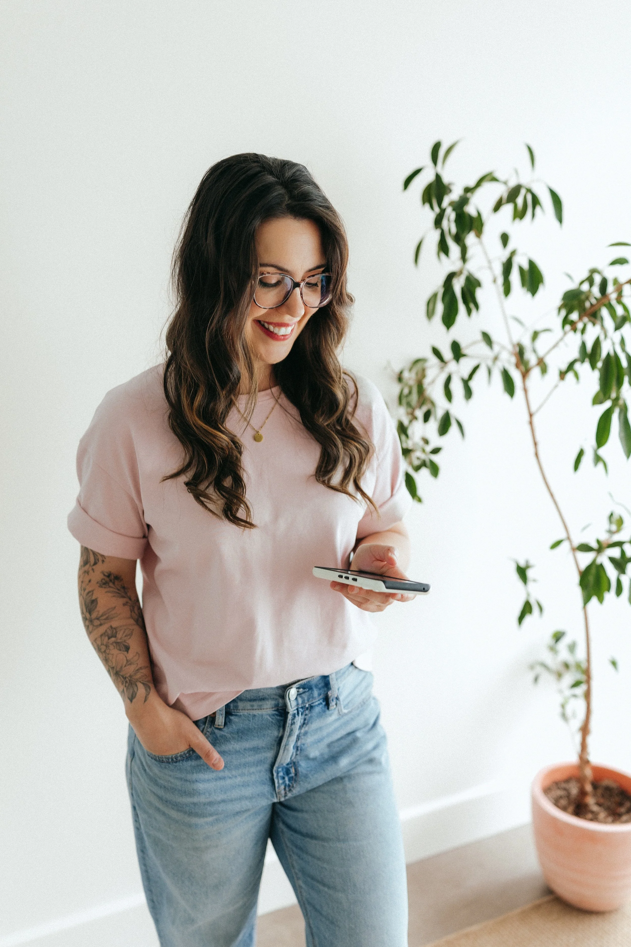 Une femme souriante qui tient son cellulaire, portant un t-shirt rose et un jean, devant un fond beige avec une plante derrière.