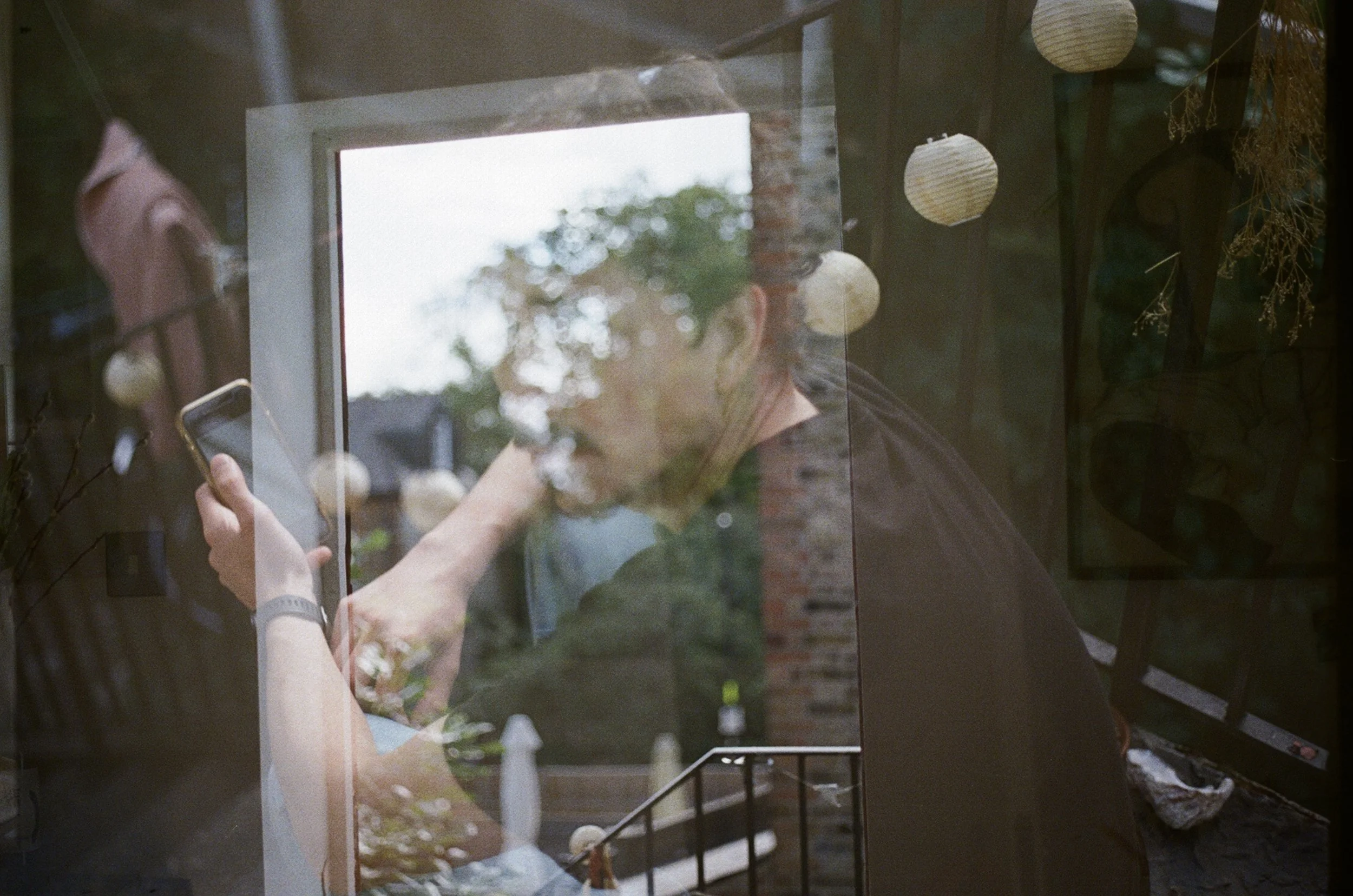 A person is seen through a window, holding a phone with outdoor scenery reflected in the glass, including trees and a cloudy sky.
