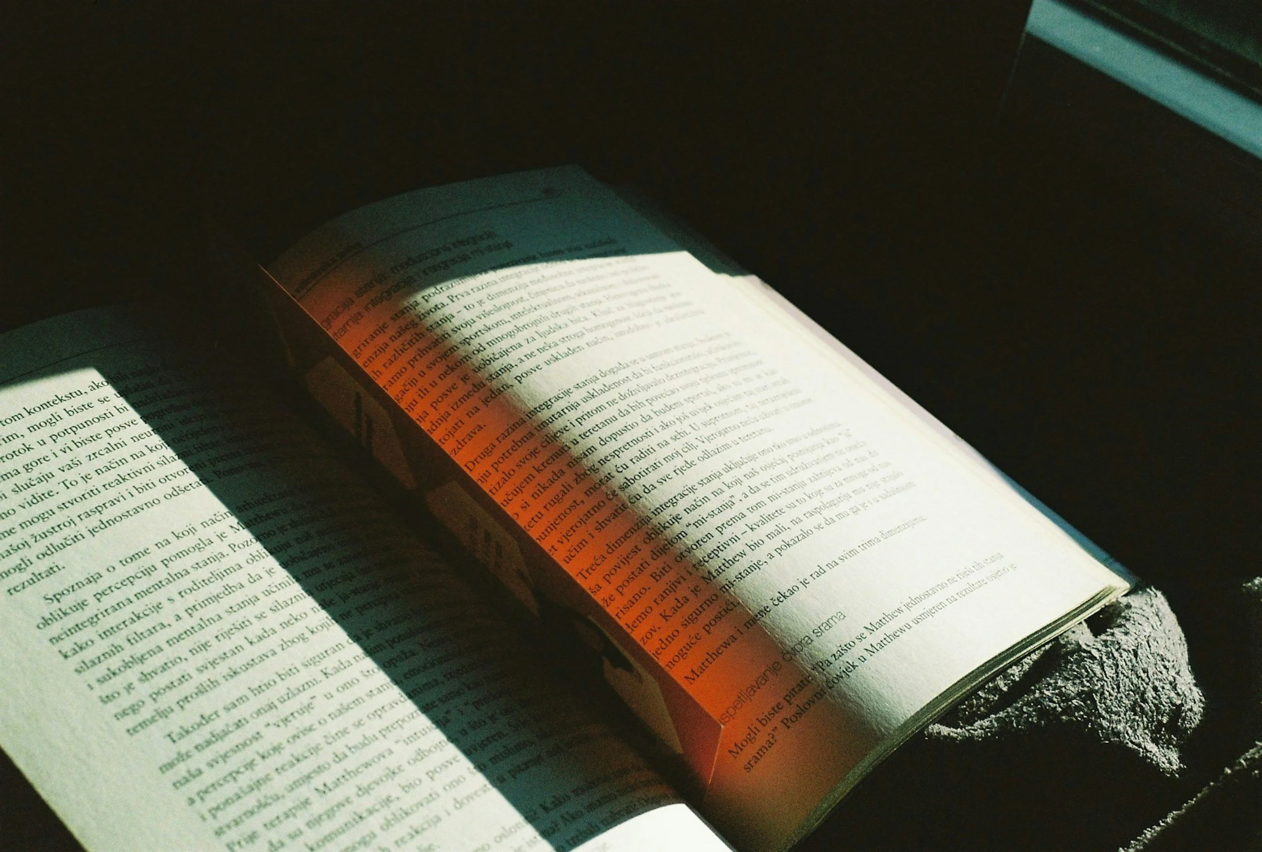 Åpen bok med tekst og en oransje markering, plassert inntil et vindu med sollys som skaper skygger på sidene.