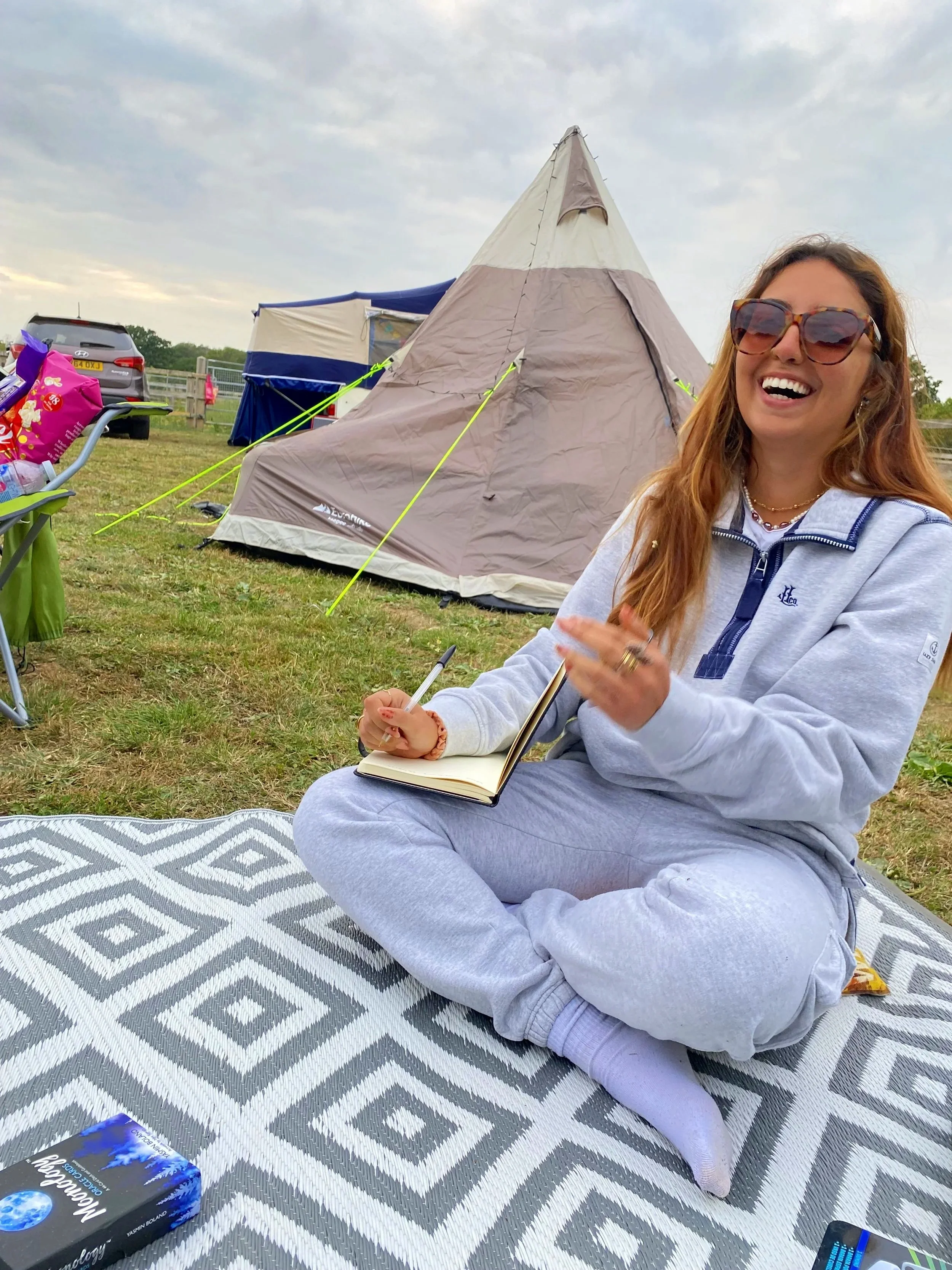 A woman sitting cross-legged on a patterned outdoor mat, smiling, with a notebook and pen, in front of a beige tent with a sky overhead.