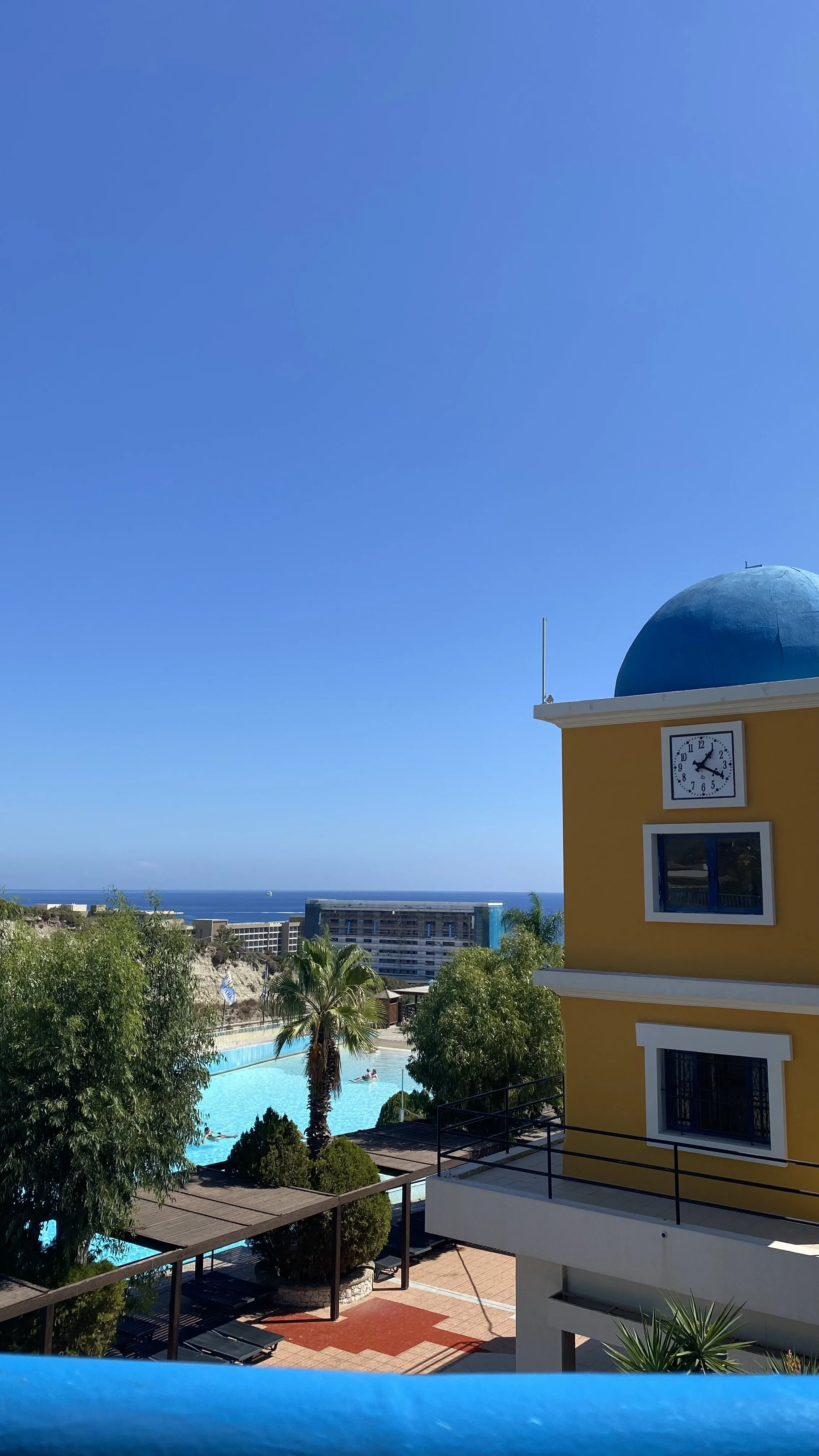 View of a bright blue sky over a coastal hotel, with a yellow building featuring a blue dome and a clock, a swimming pool, palm trees, and other hotel buildings in the background.