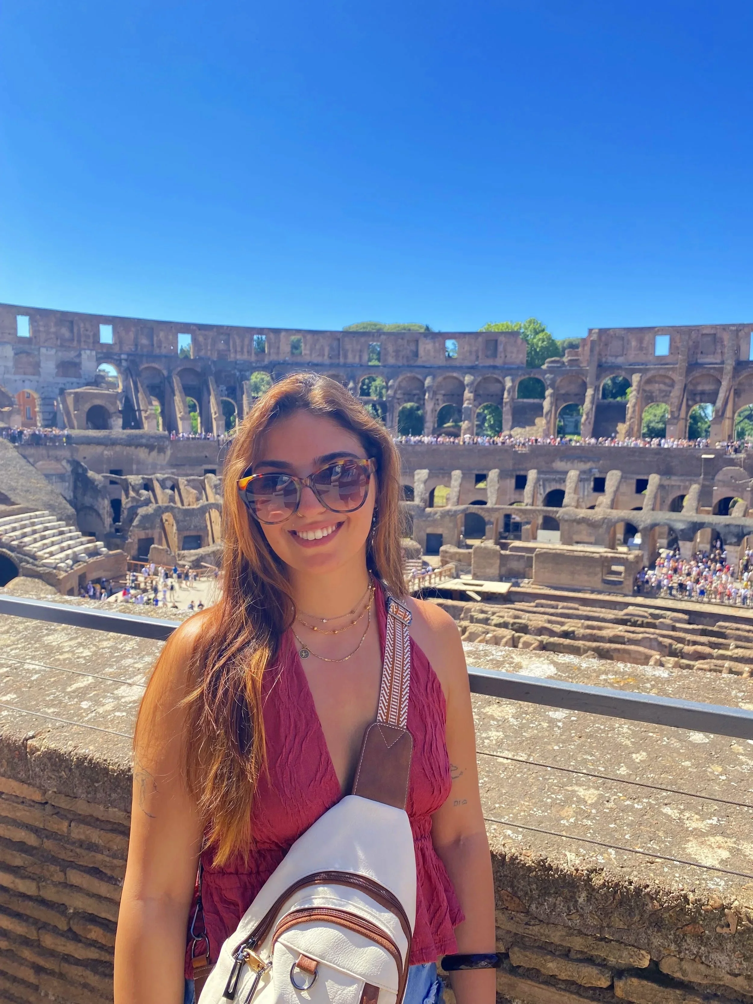 A woman with long hair, wearing sunglasses, a red sleeveless top, and layered necklaces, stands in front of the ancient Roman Colosseum on a sunny day.