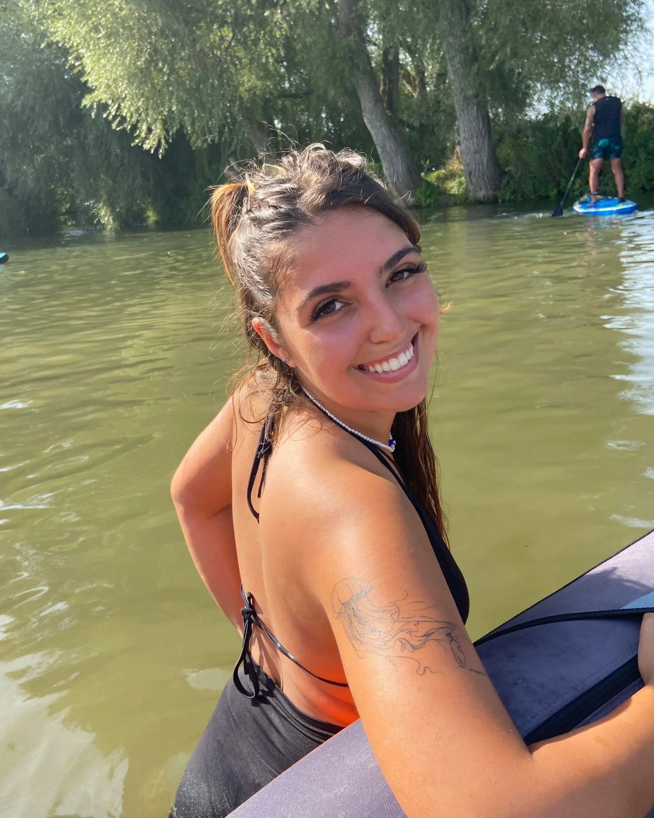 Smiling young woman with tattoo on arm floating in a body of water near trees, with paddleboarder in the background.