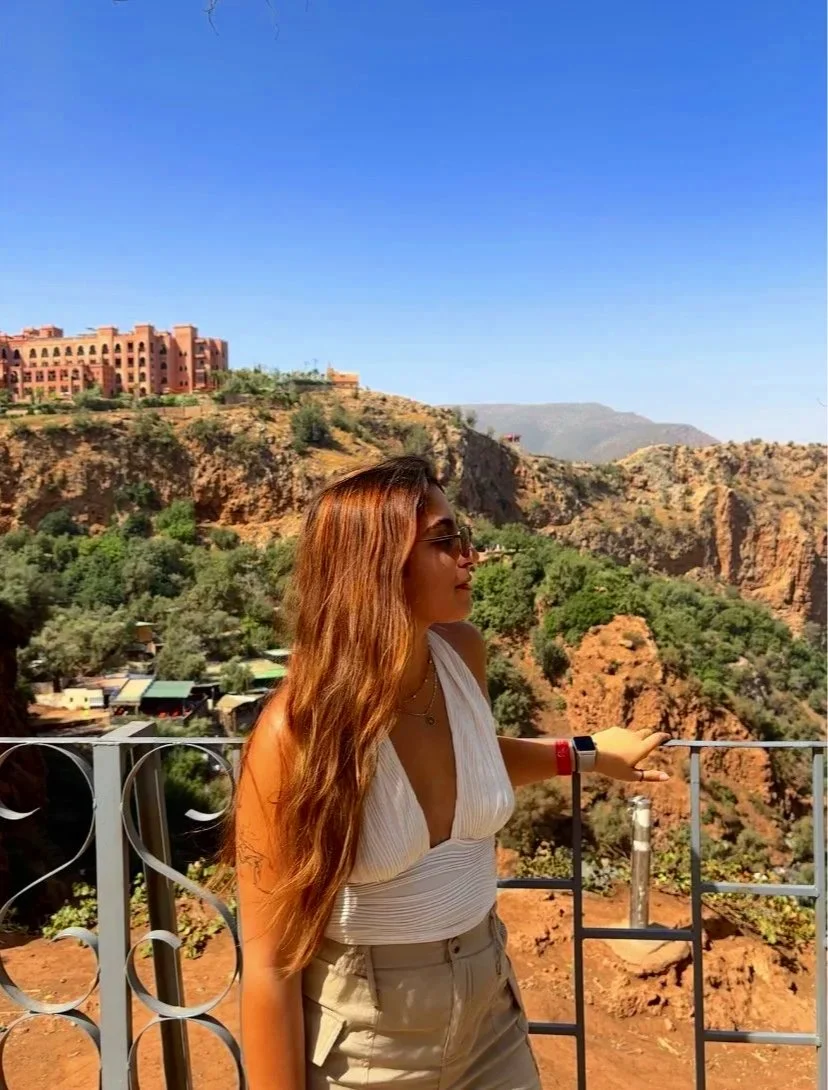 A woman with long wavy hair, wearing sunglasses and a sleeveless white top, standing near a metal railing overlooking a canyon with trees and rocky cliffs. A building is visible on the hill in the background under a clear blue sky.