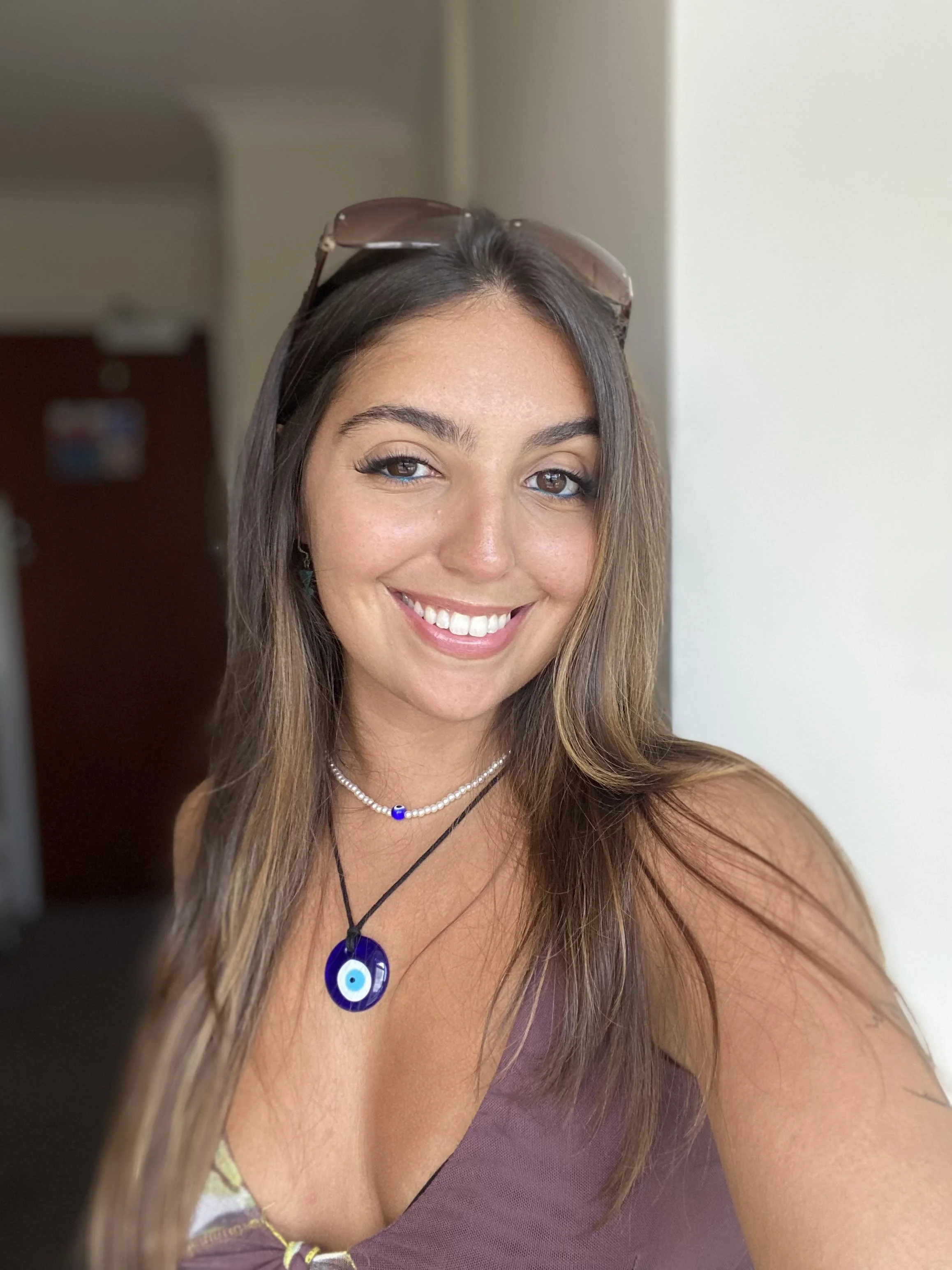 A young woman with long brown hair, smiling at the camera, wearing a purple top and layered necklaces, with sunglasses on her head.