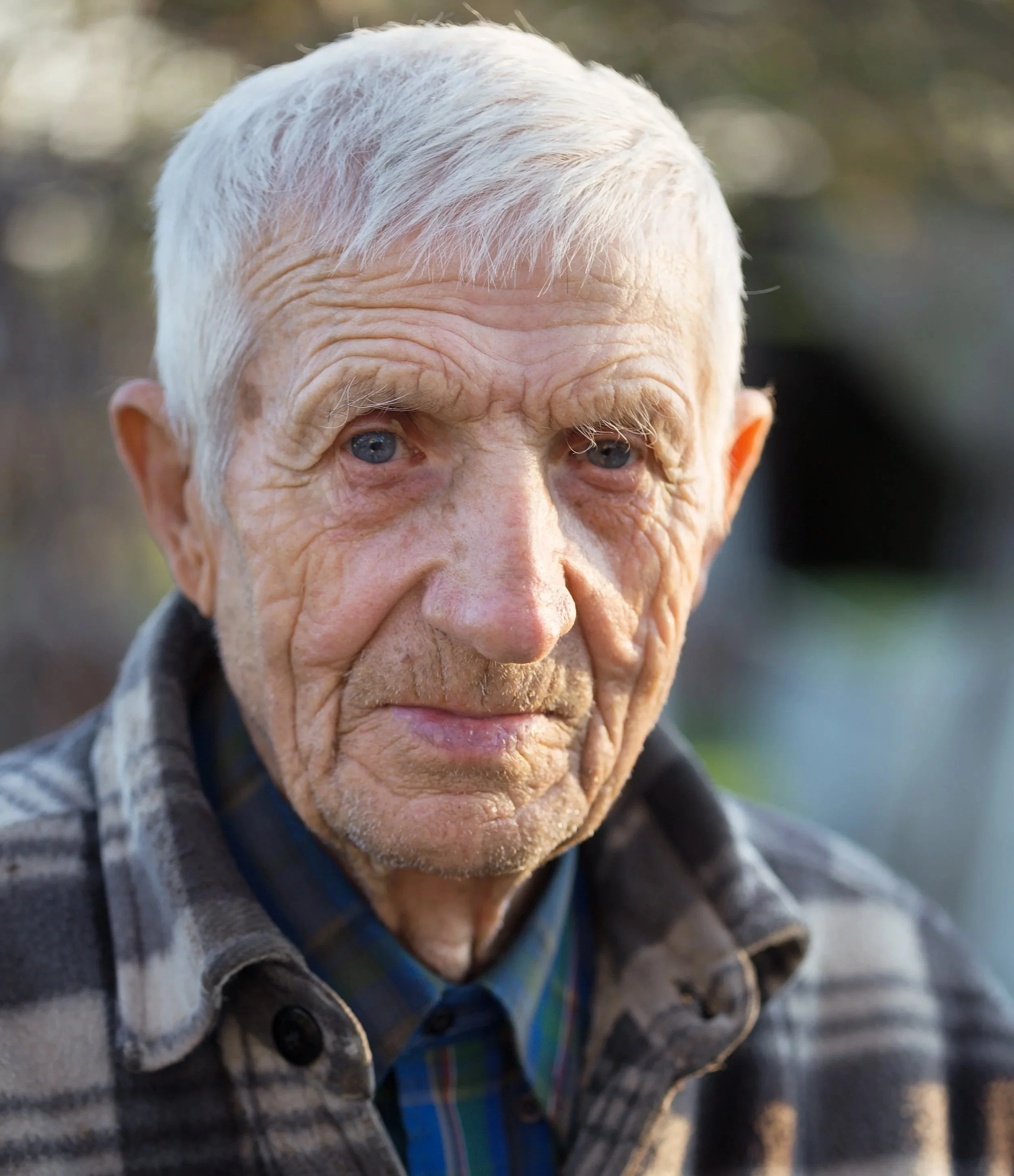 Nahaufnahme eines alten Mannes mit grauem Haar, blauen Augen, Falten im Gesicht und einem ernsten Ausdruck, trägt eine kariert Flanelljacke und einen Hemdkragen, unscharfer Hintergrund im Freien.