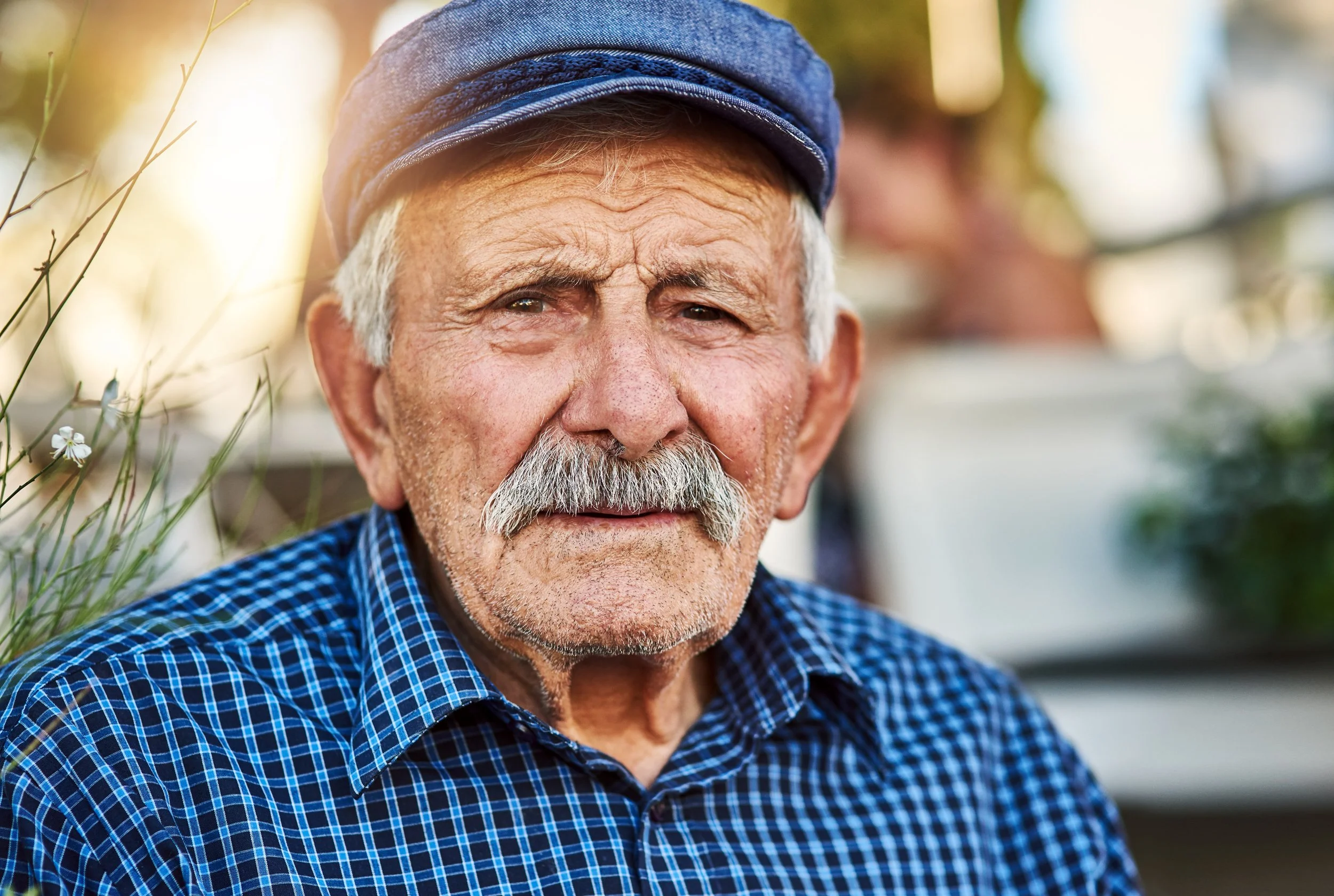 Porträt eines älteren Mannes mit weißem Schnurrbart, trägt eine blaue Kappe und ein blau-kariertes Hemd, sitzt im Freien bei Sonnenuntergang.