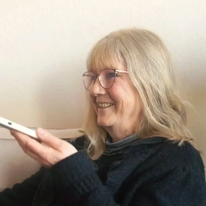 Eine ältere Frau mit Brille hält ein Smartphone und lächelt.